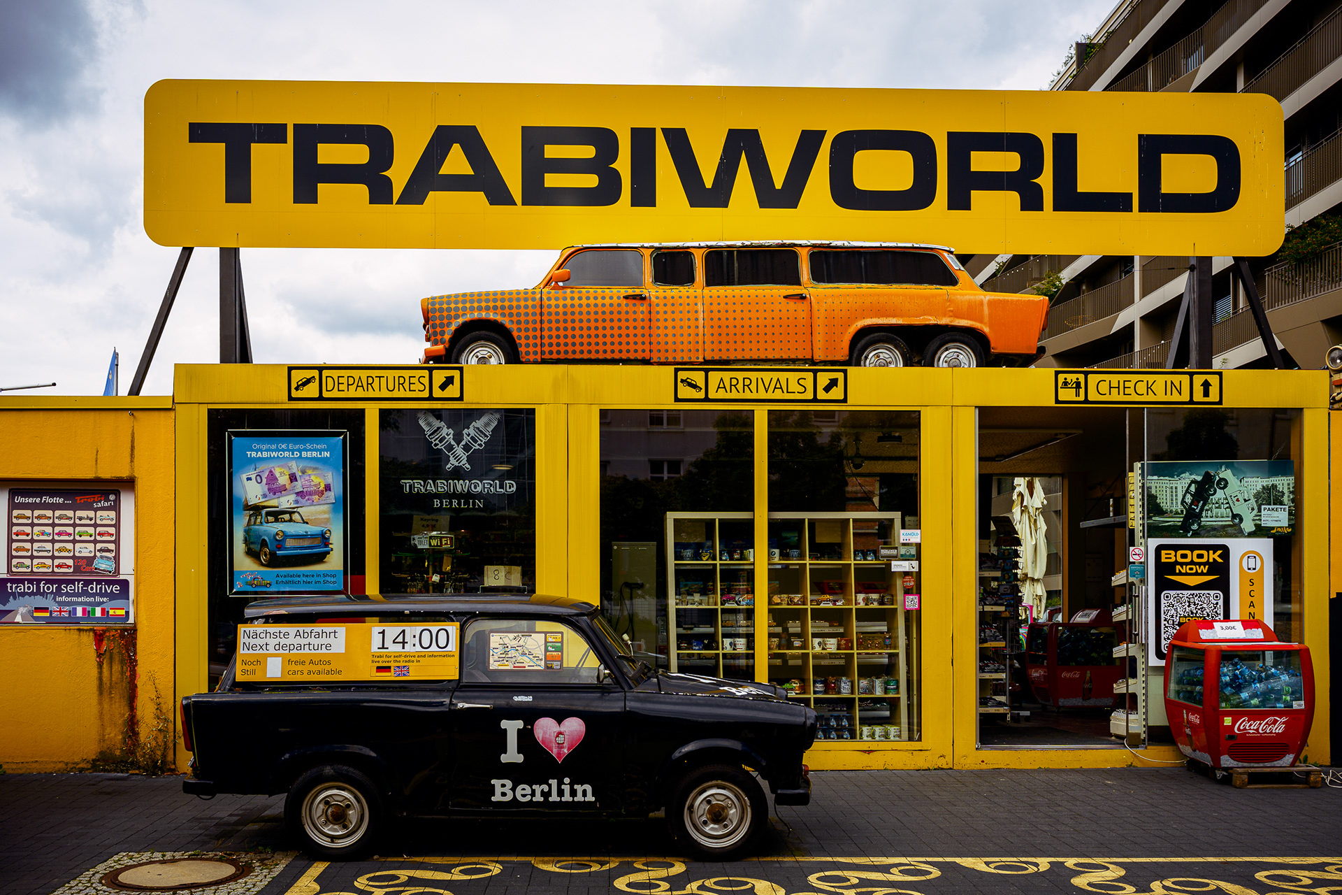 Vibrant photo of "Trabi World" in Berlin. A bright yellow industrial structure supports an orange spotted Trabant limousine on the roof. Below, a black Trabant and a red Coca-Cola cooler sit on the street level. The colors are saturated and pop-art style.