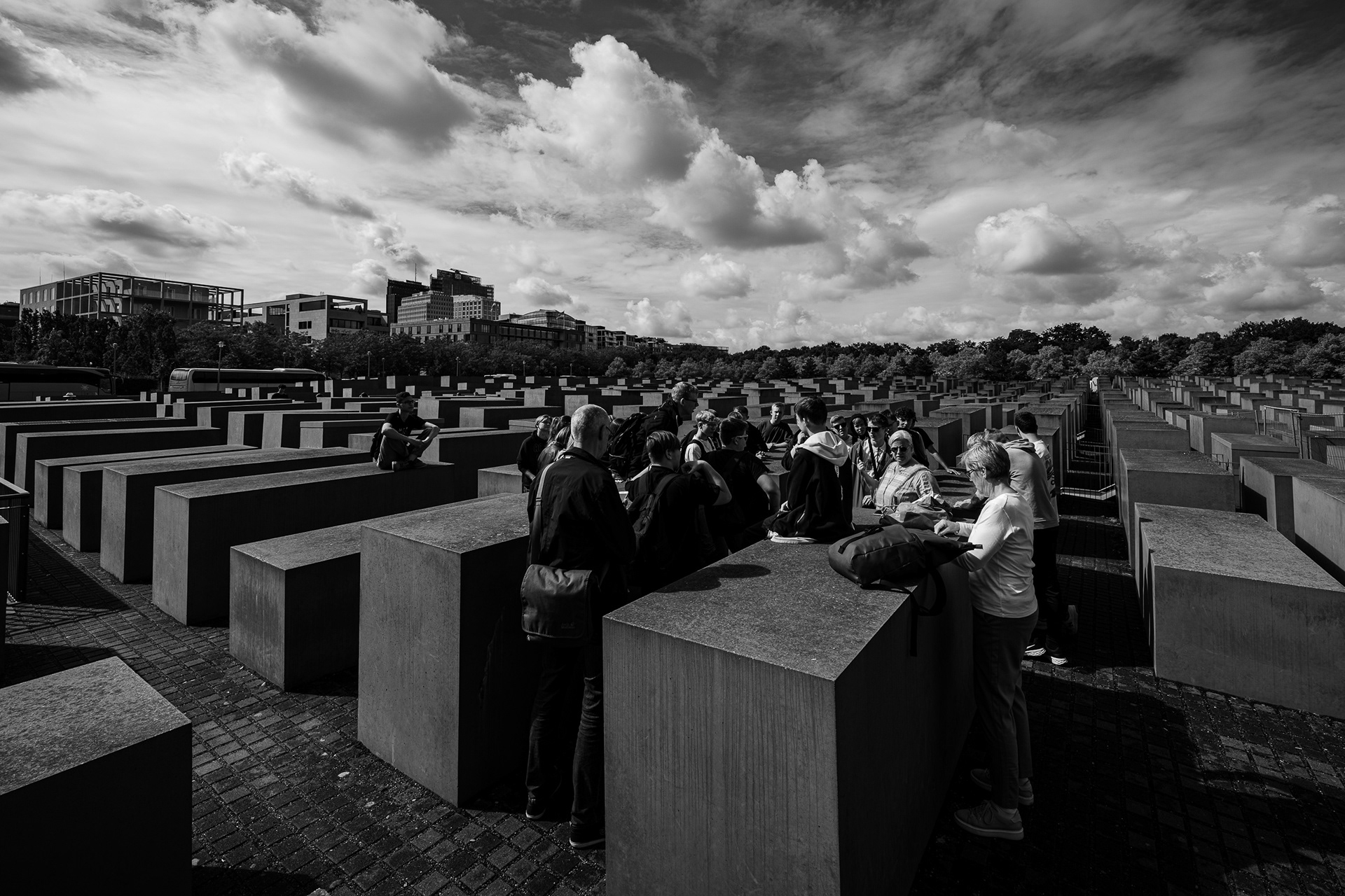 Black and white photograph of the Memorial to the Murdered Jews of Europe. A group of visitors sits and stands on the dark concrete stelae, providing scale to the vast grid. The sky is dramatic and moody, contrasting with the geometric shadows. Caption: