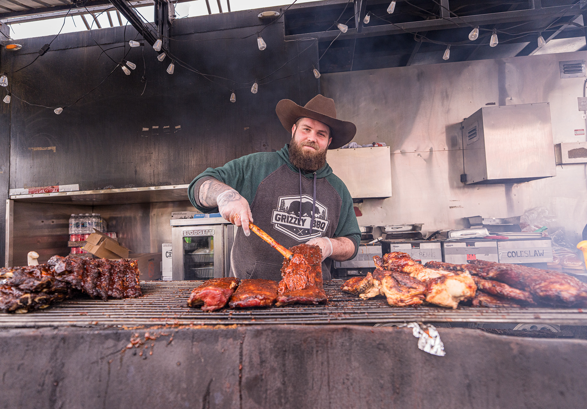 A bearded pitmaster in a cowboy hat preparing ribs at the Grizzly BBQ stall during the Cloverdale Rodeo.
