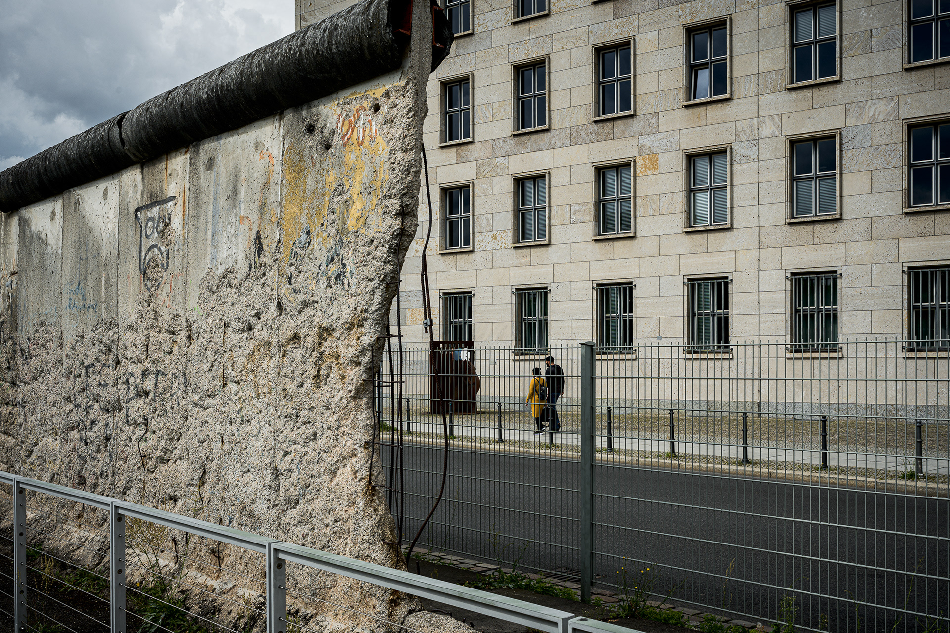 Street photograph of a section of the Berlin Wall. The rough, pockmarked concrete is covered in faint graffiti. A pedestrian wearing a bright yellow coat walks past, providing a sharp color contrast against the grey texture of the former border.