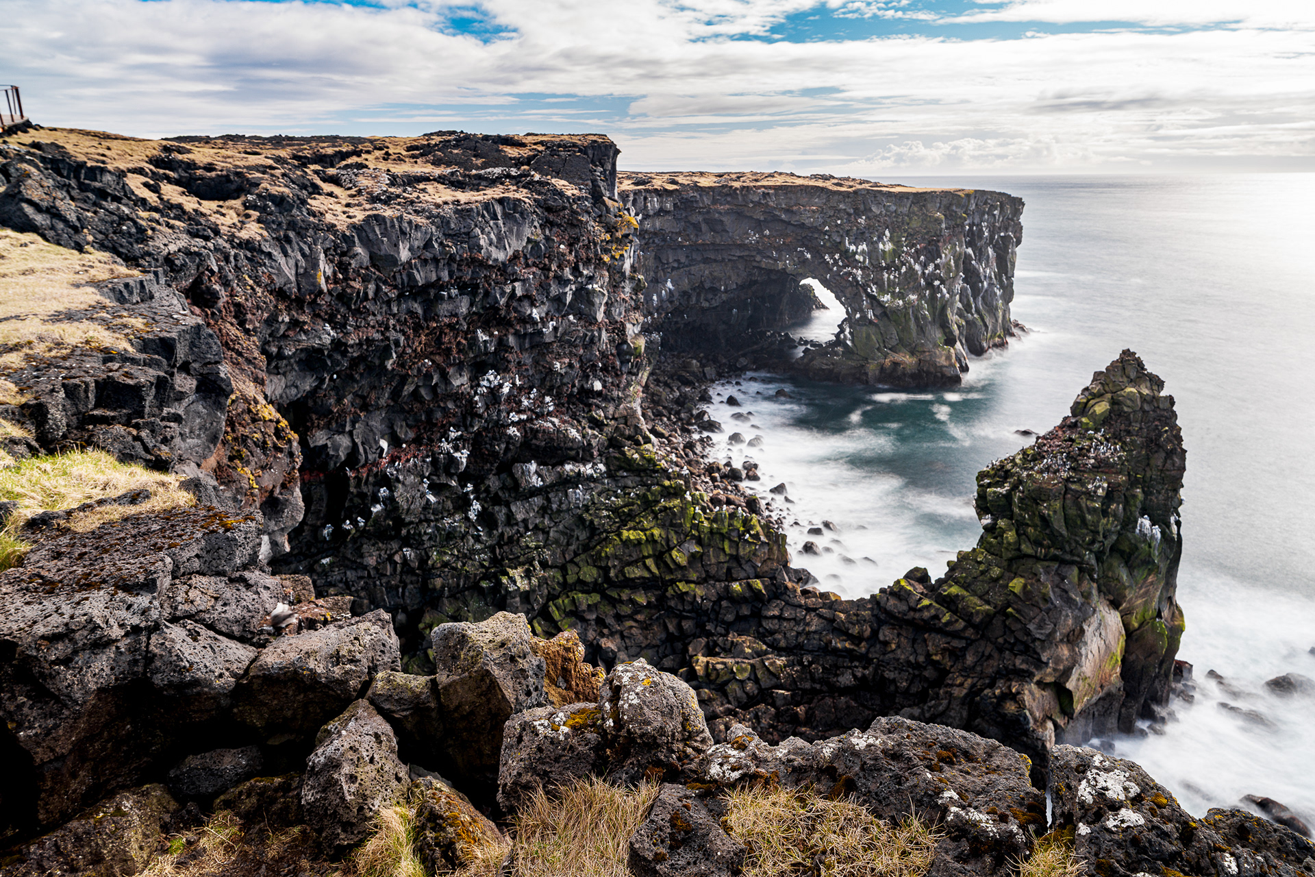 Wide-angle view of a rugged volcanic rock arch and sea stack on the Icelandic coastline, with white waves crashing against the dark basalt cliffs.