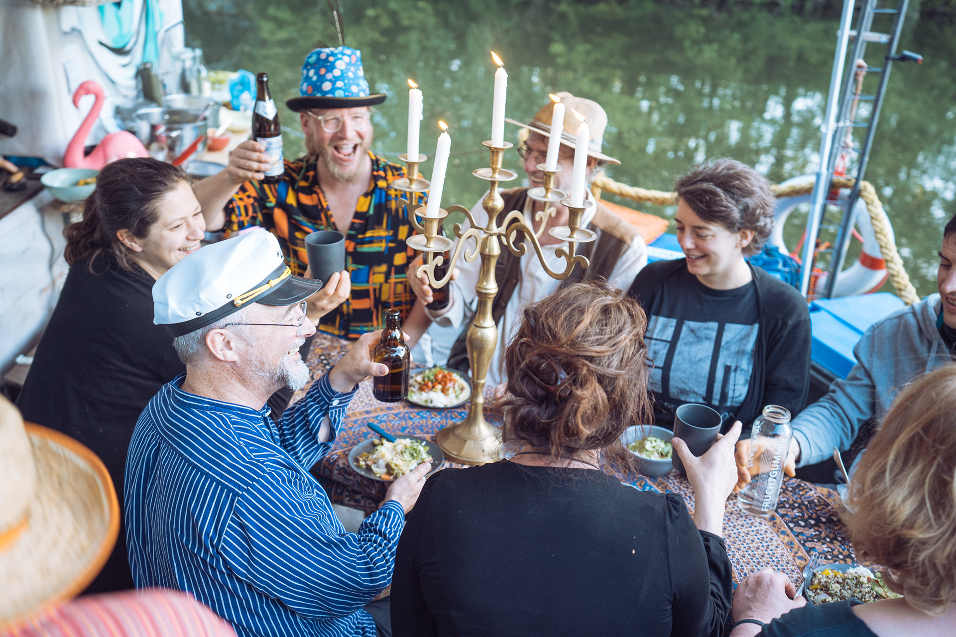 Auf dem Foto sitzen rund um um einen Tisch viele Menschen zusammen, die gerade mit ihren Getränken anstoßen. Im Hintergrund ist das Wasser eines Flusses zu sehen.