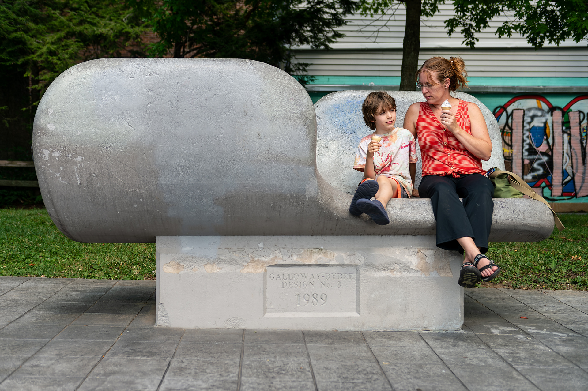 Kim Wright and her son Felix Wright, 8, of Bloomfield, Ind., eat ice cream atop the Bench in the Shape of Opposing Wings limestone sculpture at Peoples Park, Wednesday, July 10, 2024, in Bloomington, Ind. The Wrights have a tradition of visiting Bloomington to see the library and enjoy ice cream in the park. (Vincent Hsiao/HSJI)