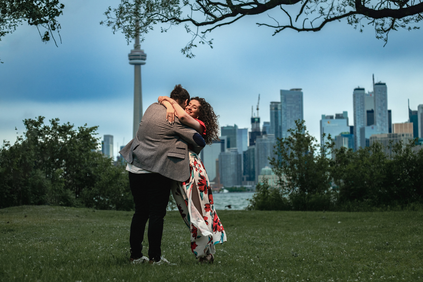 Couple hugging under tree branches with the Toronto skyline and CN Tower in the background.