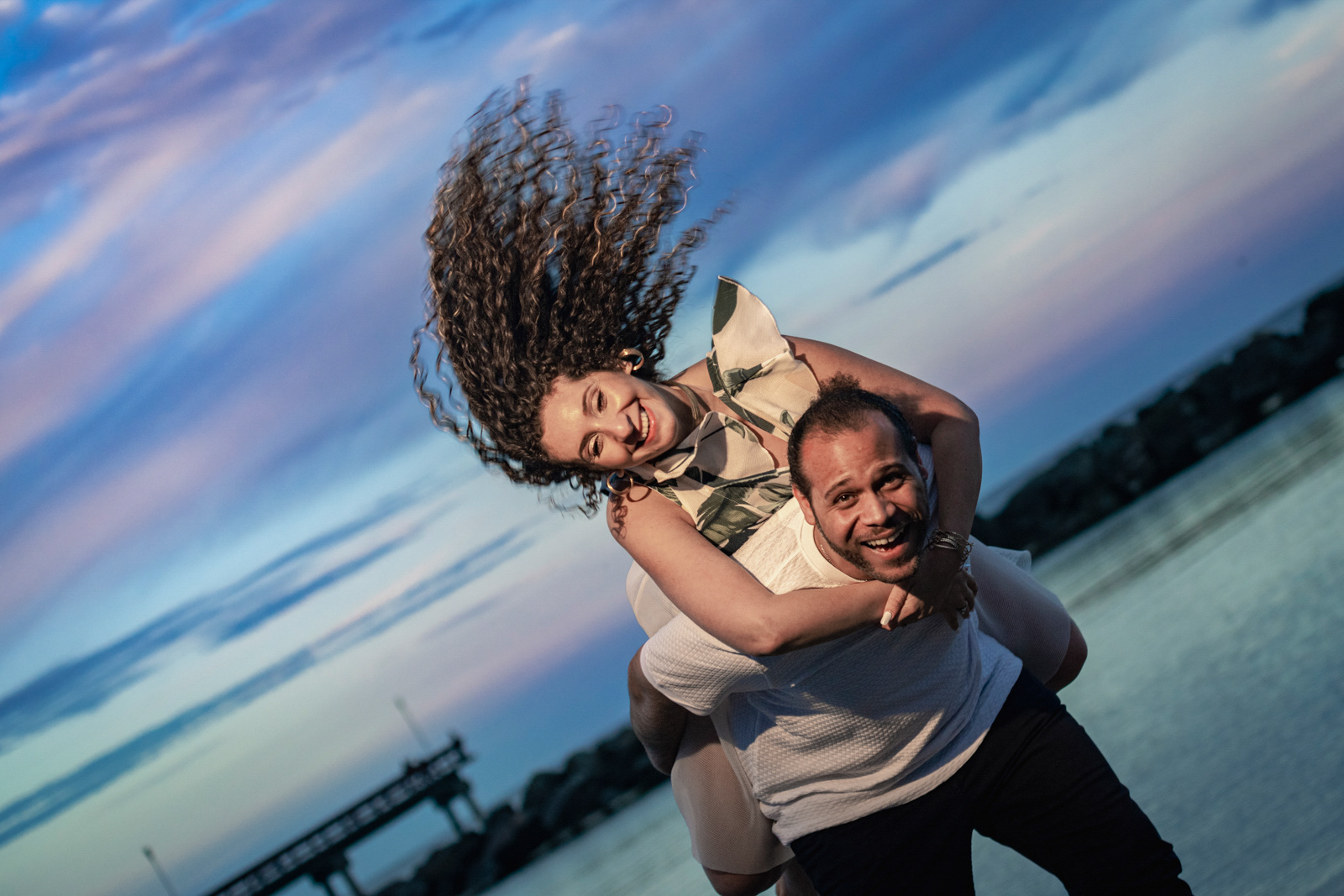 Bride-to-be’s curly hair flying as she laughs during playful piggyback ride at the lake, pastel evening sky.