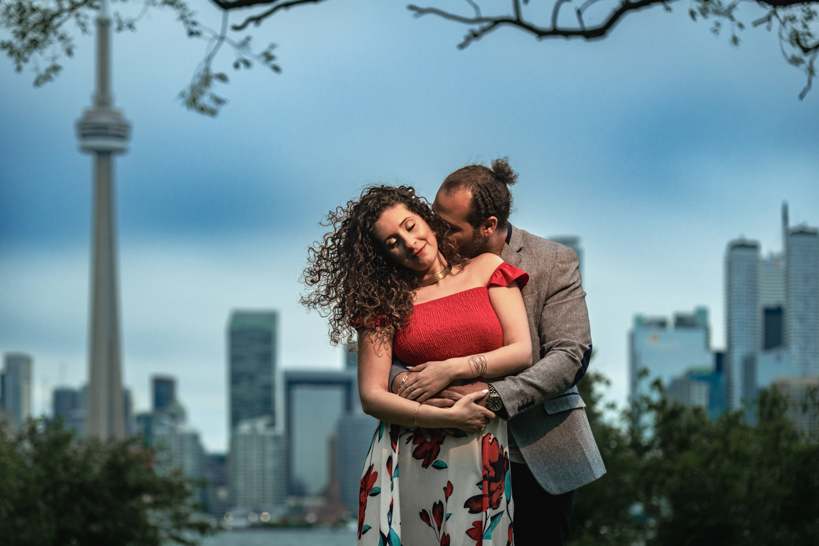 Intimate engagement photo of a couple in front of the Toronto skyline at sunset, framed by trees with the CN Tower in the background.