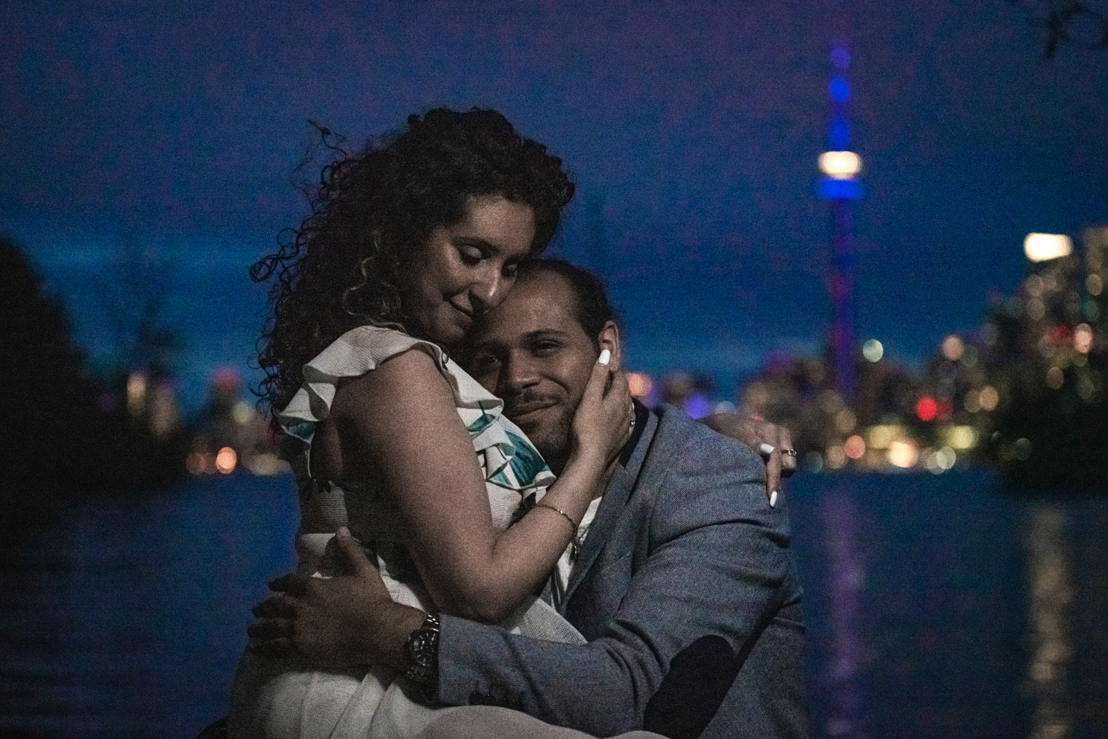Romantic couple cuddling during their Toronto engagement session, CN Tower lights glowing in the background.