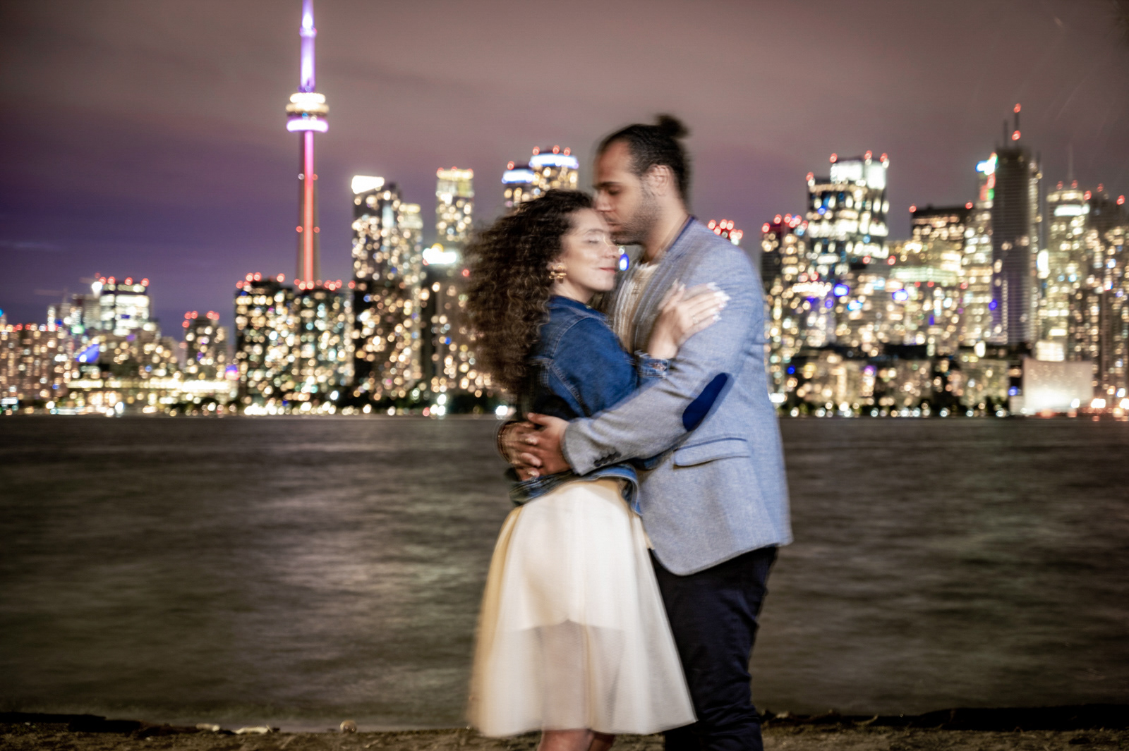 Romantic engagement photo of a couple embracing in front of the Toronto skyline at night, with the CN Tower glowing in vibrant lights.