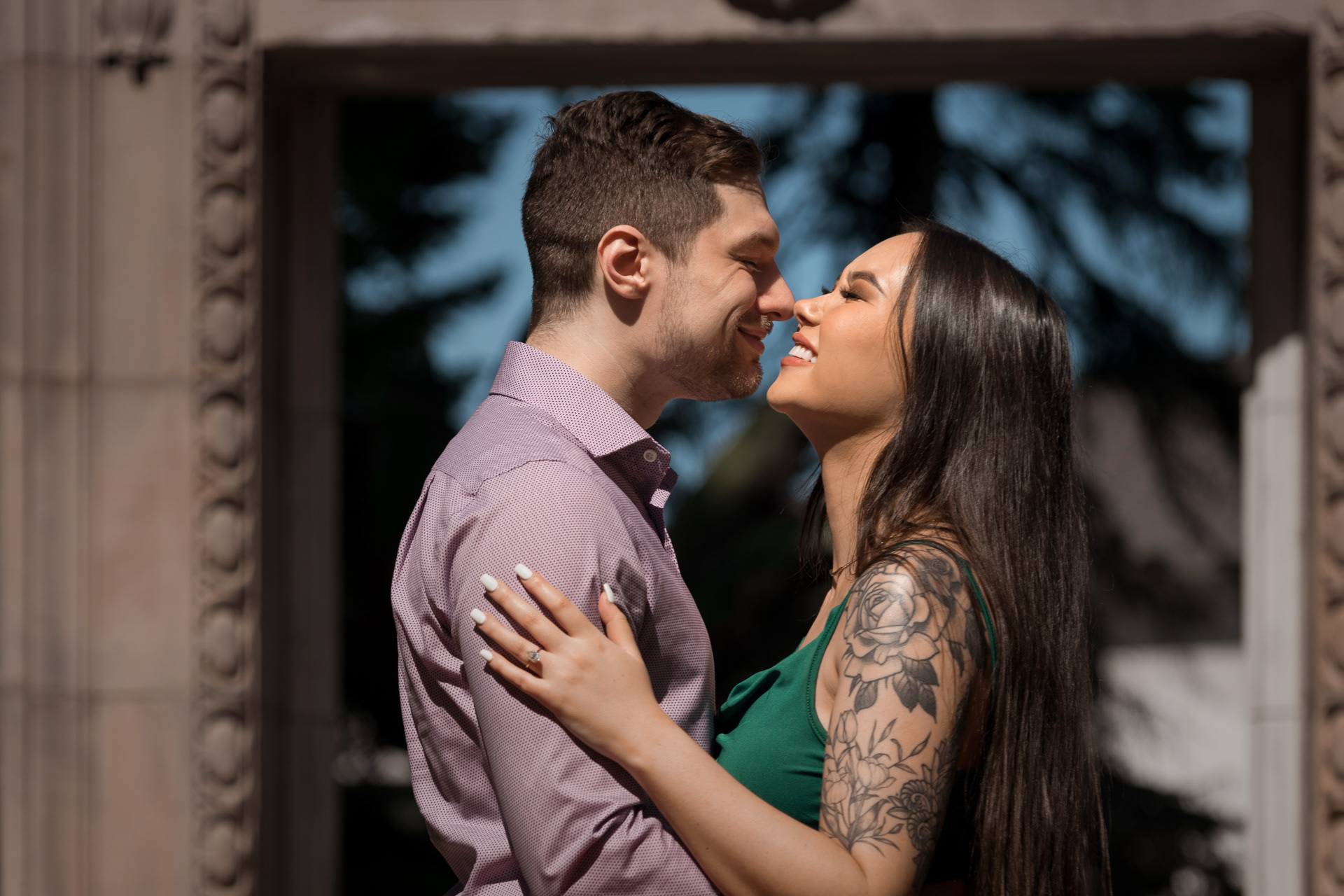 Couple smiling nose-to-nose, showing off engagement ring in front of Guild Park archway.