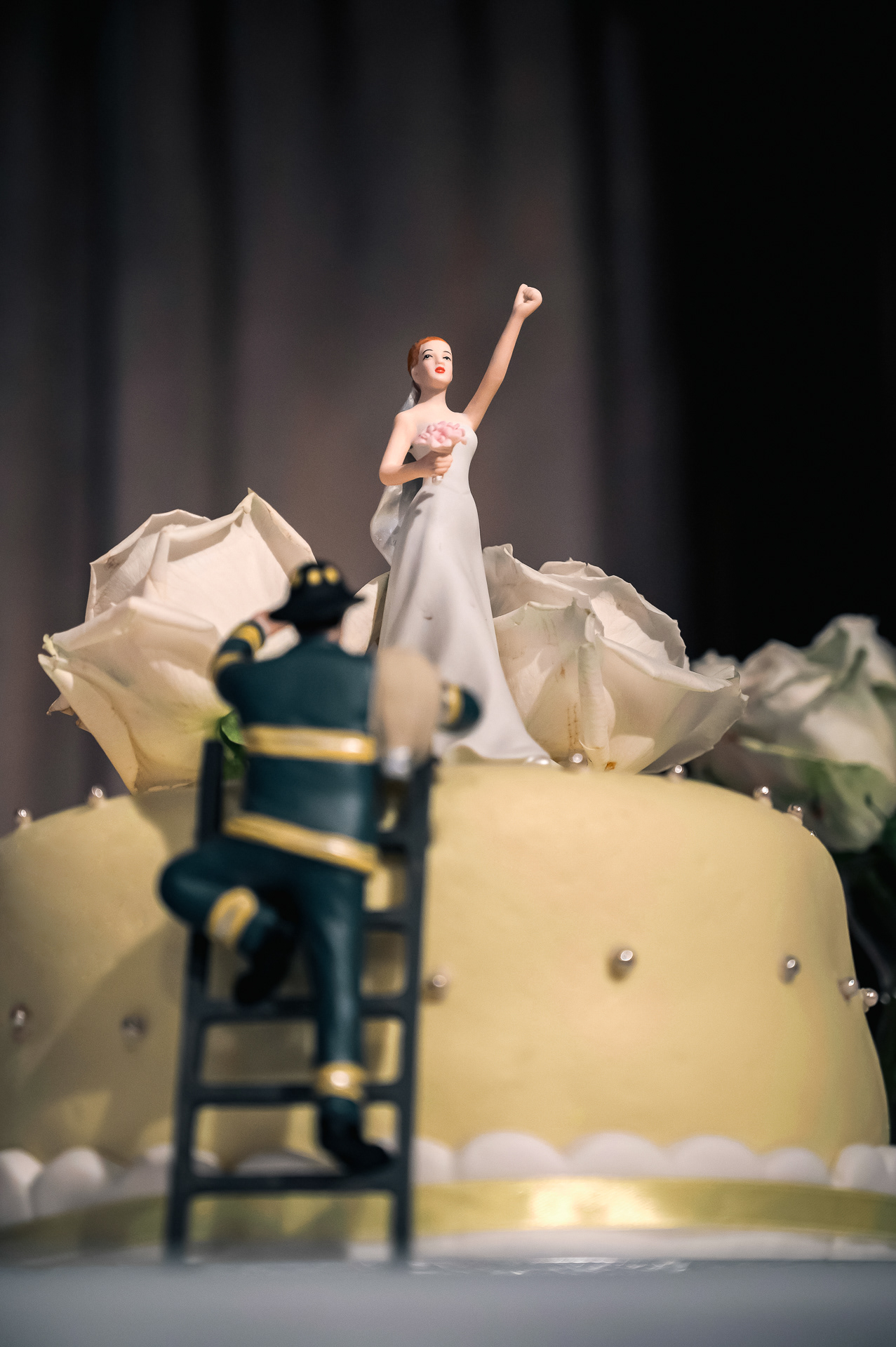 Playful cake topper showing a miniature groom fireman climbing a ladder to reach his bride.