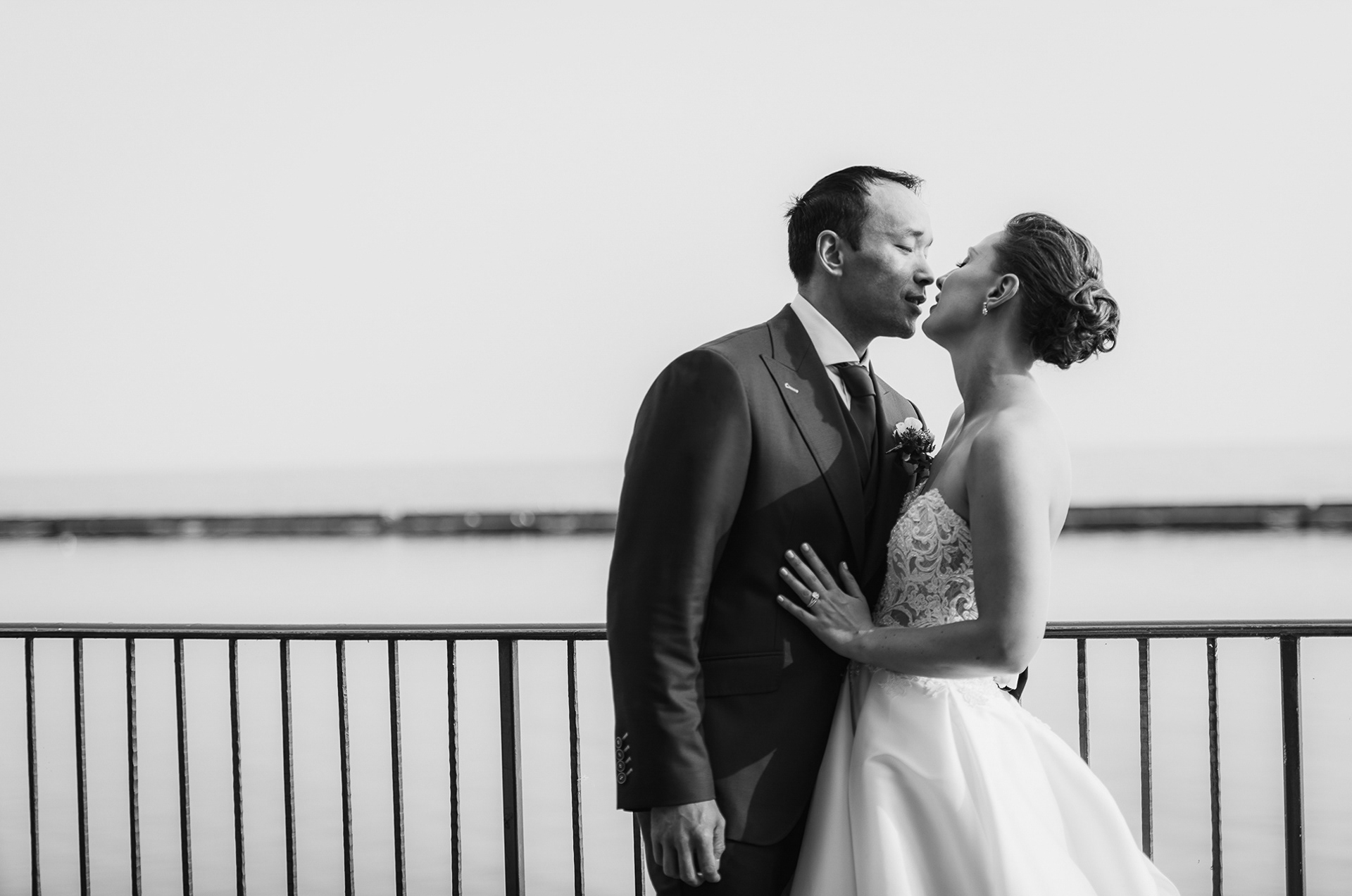 Black and white portrait of the couple kissing by the lakeshore railing.