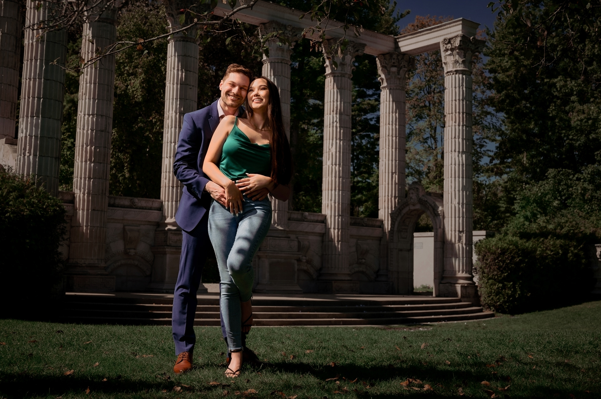 Couple embracing in front of the stone columns at Guild Park, with the groom-to-be smiling behind his fiancée.