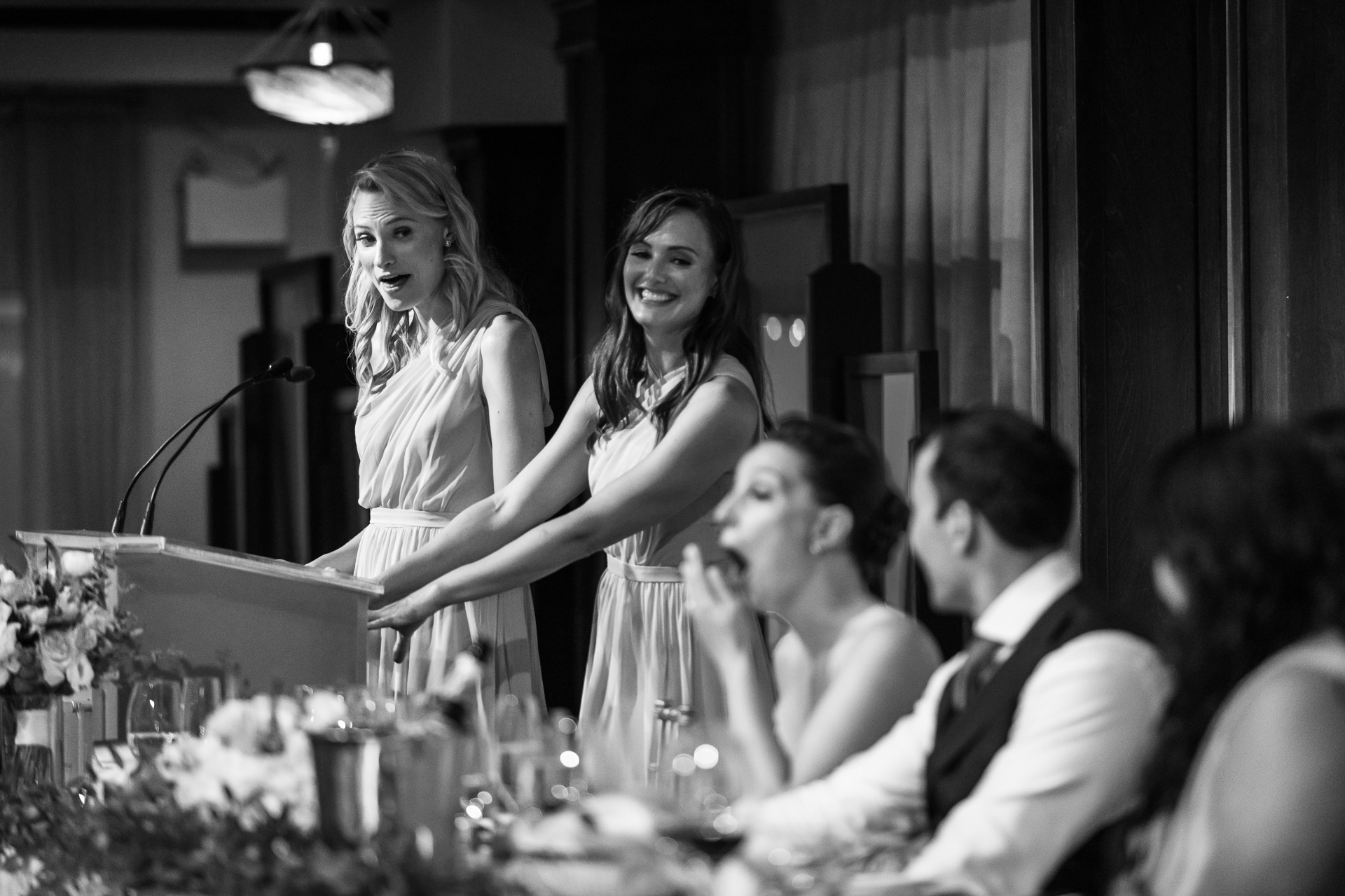 The bride, slightly blurred in the foreground, bursts into a shocked laugh while listening to her bridesmaids’ speeches during the reception at Palais Royale.