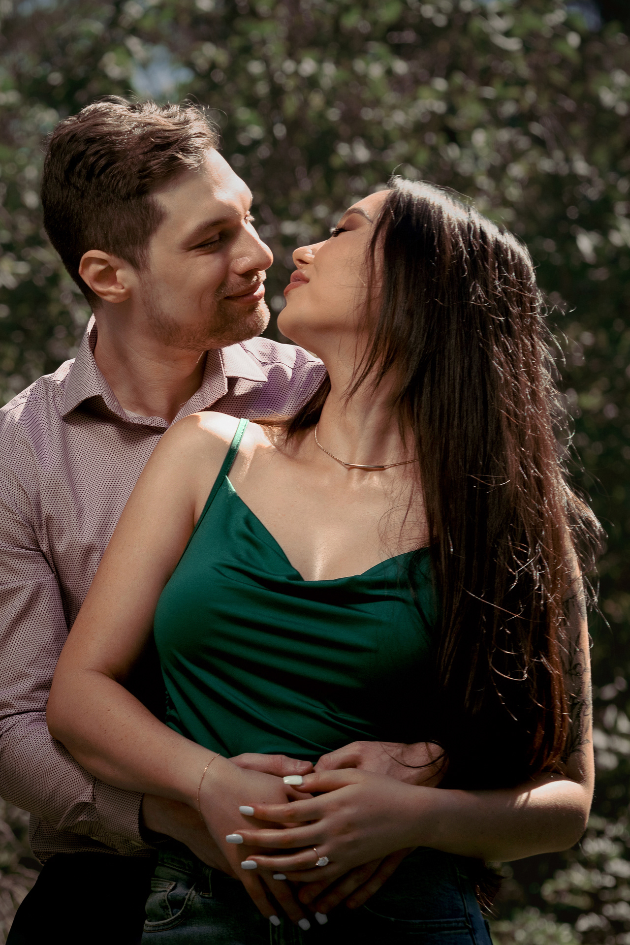 Groom-to-be holding his fiancée close, almost kissing under soft sunlight in the trees.