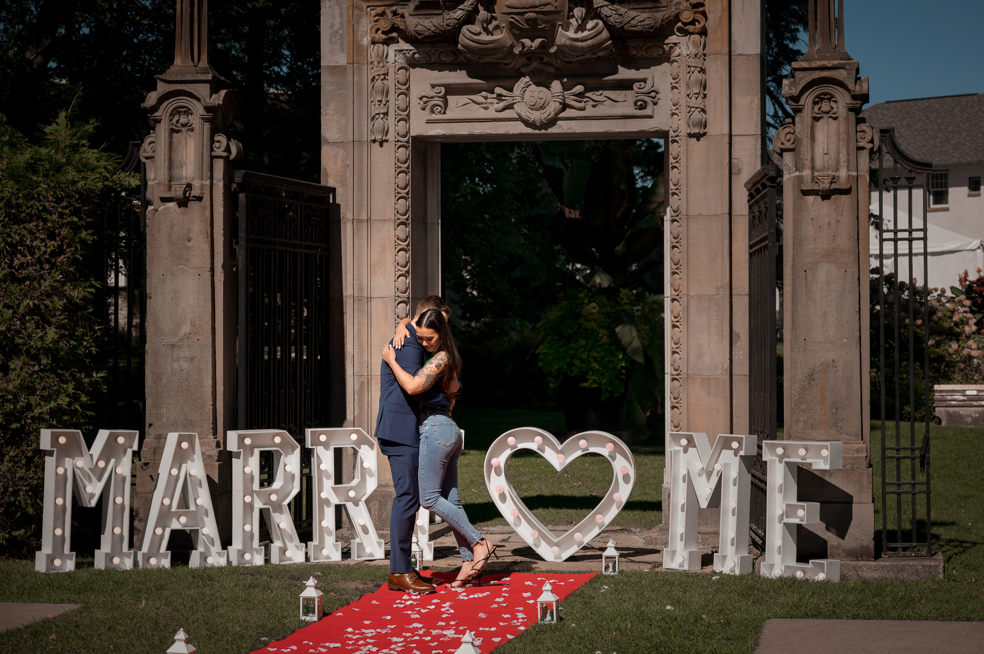 Groom-to-be embracing fiancée in front of a “Marry Me” sign under a historic stone archway at Guild Park.