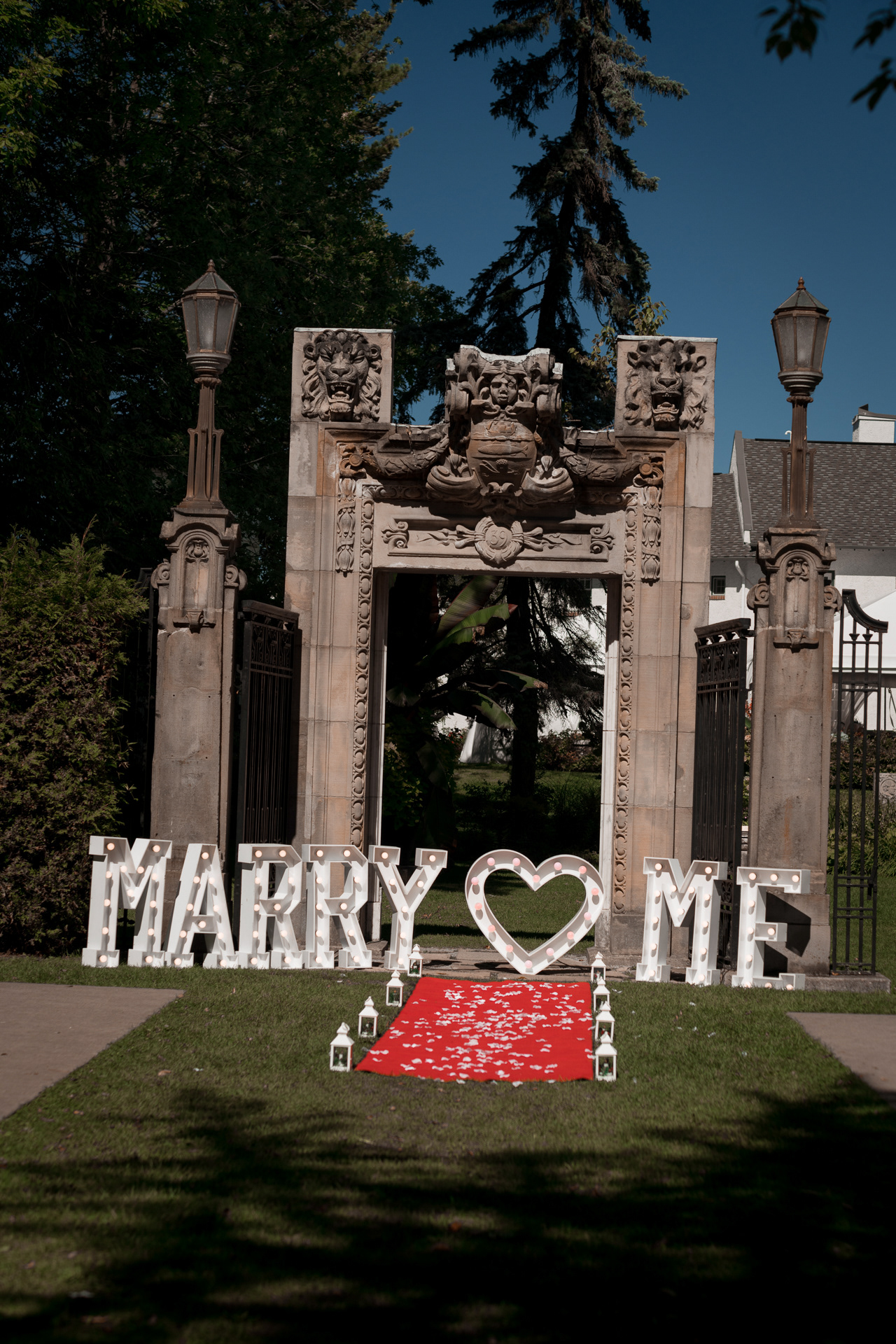 Proposal setup with large light-up “Marry Me” letters, red carpet and lanterns in front of the archway.