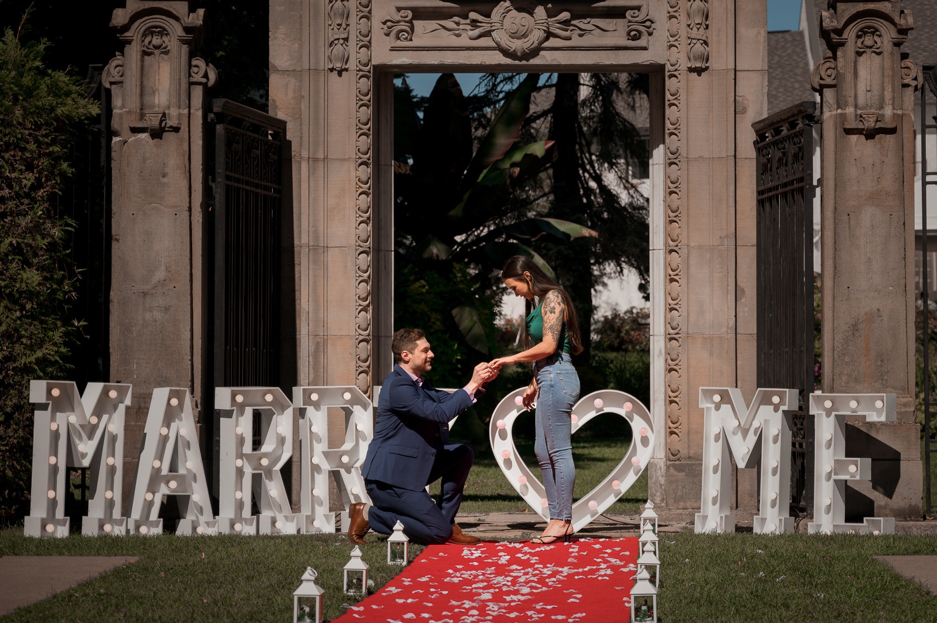 Groom-to-be kneeling on red carpet under the arch, proposing with ring in hand.