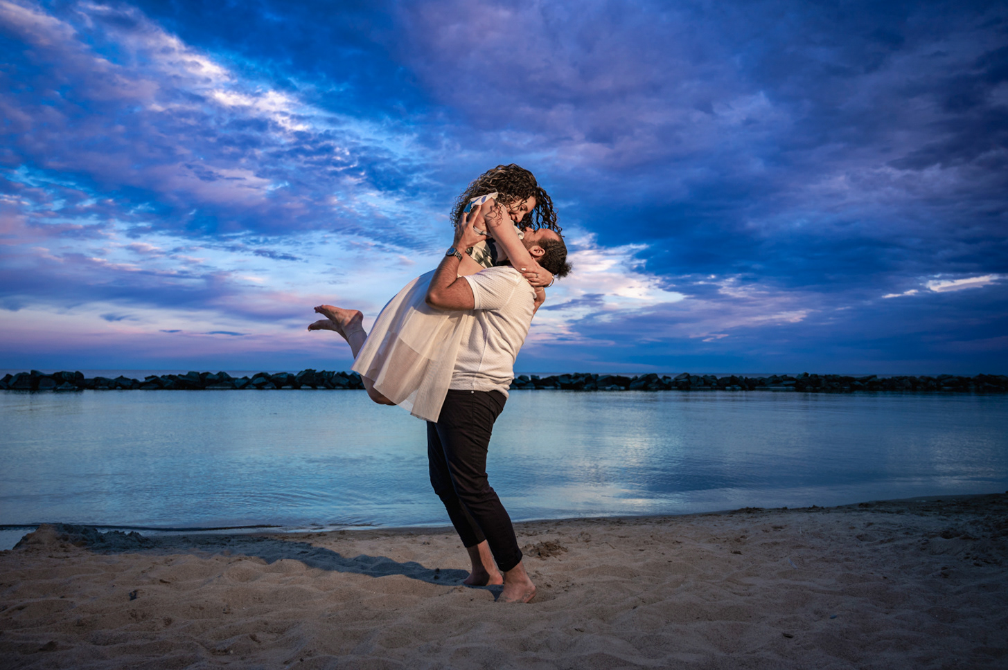 Groom-to-be lifting his fiancée joyfully on the beach at sunset, soft pastel sky over Lake Ontario.