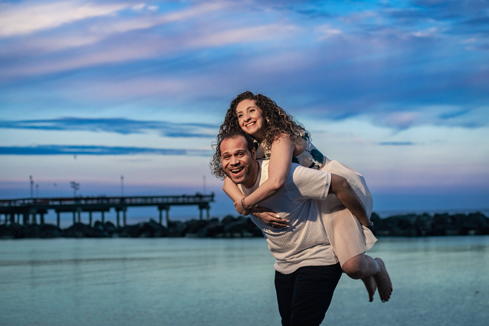 Couple laughing and playing with a piggyback ride on the beach at sunset, Toronto waterfront engagement session.