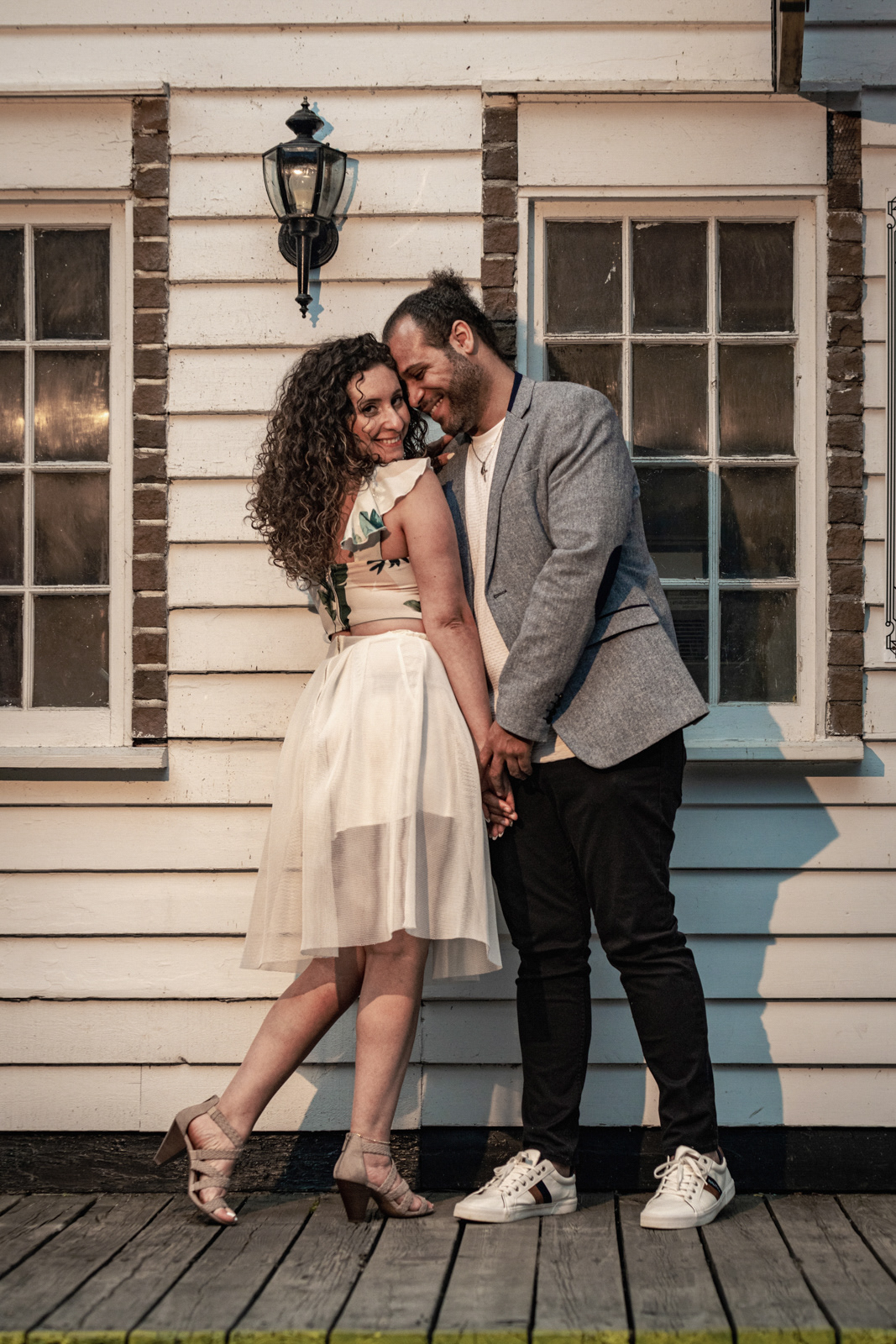 Couple leaning against a rustic white cottage wall, holding hands and smiling during Toronto island engagement session.