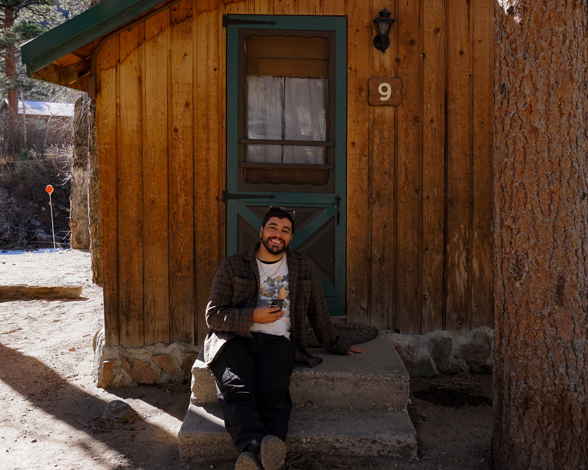 Portrait of the photographer, Gabi Cruz, sitting on a cabin step, smiling in front of a mountain stay.