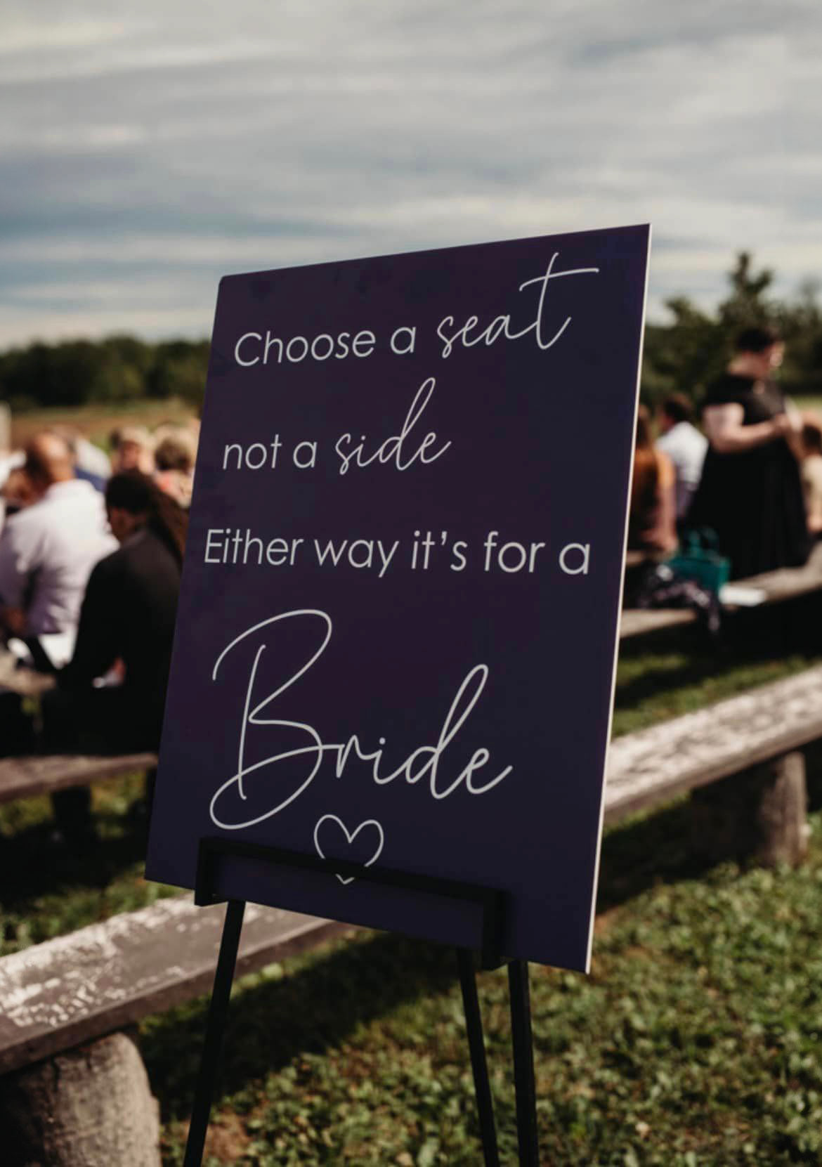A large sign that reads 'Choose a seat not a side, either way it's for a bride' leans on an easel outside a ceremony space.