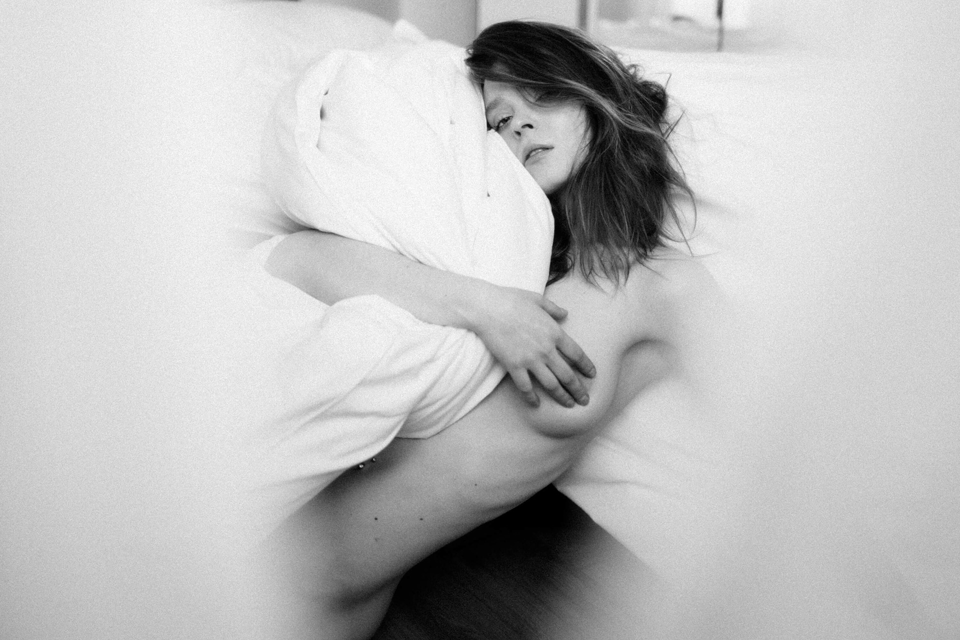 Intimate fine art nude photograph of a woman lying in bed, embracing a pillow in soft light