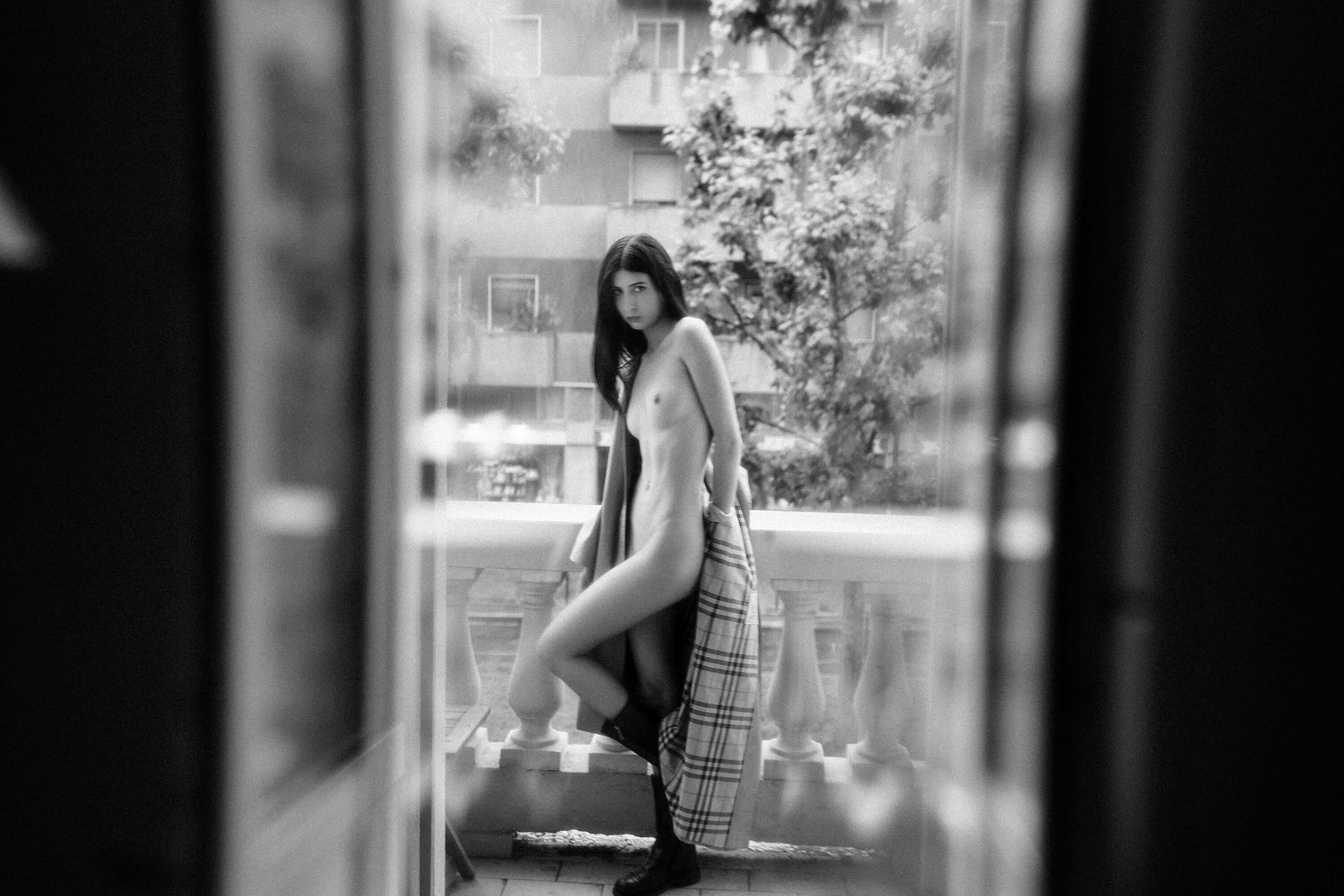 Nude woman standing on a balcony seen through a window in an urban setting