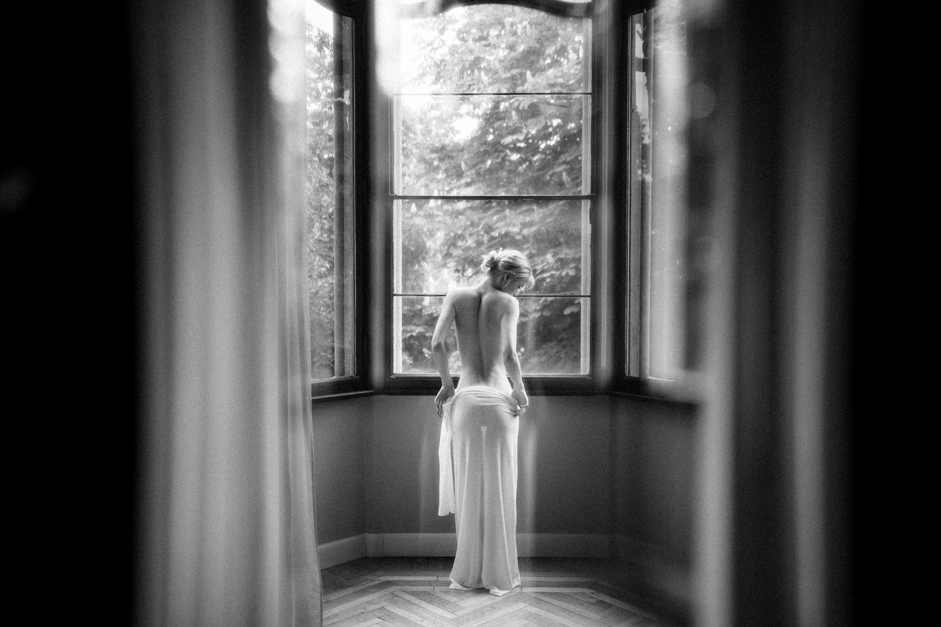 Nude woman seen from behind standing at a window with sheer fabric and natural light