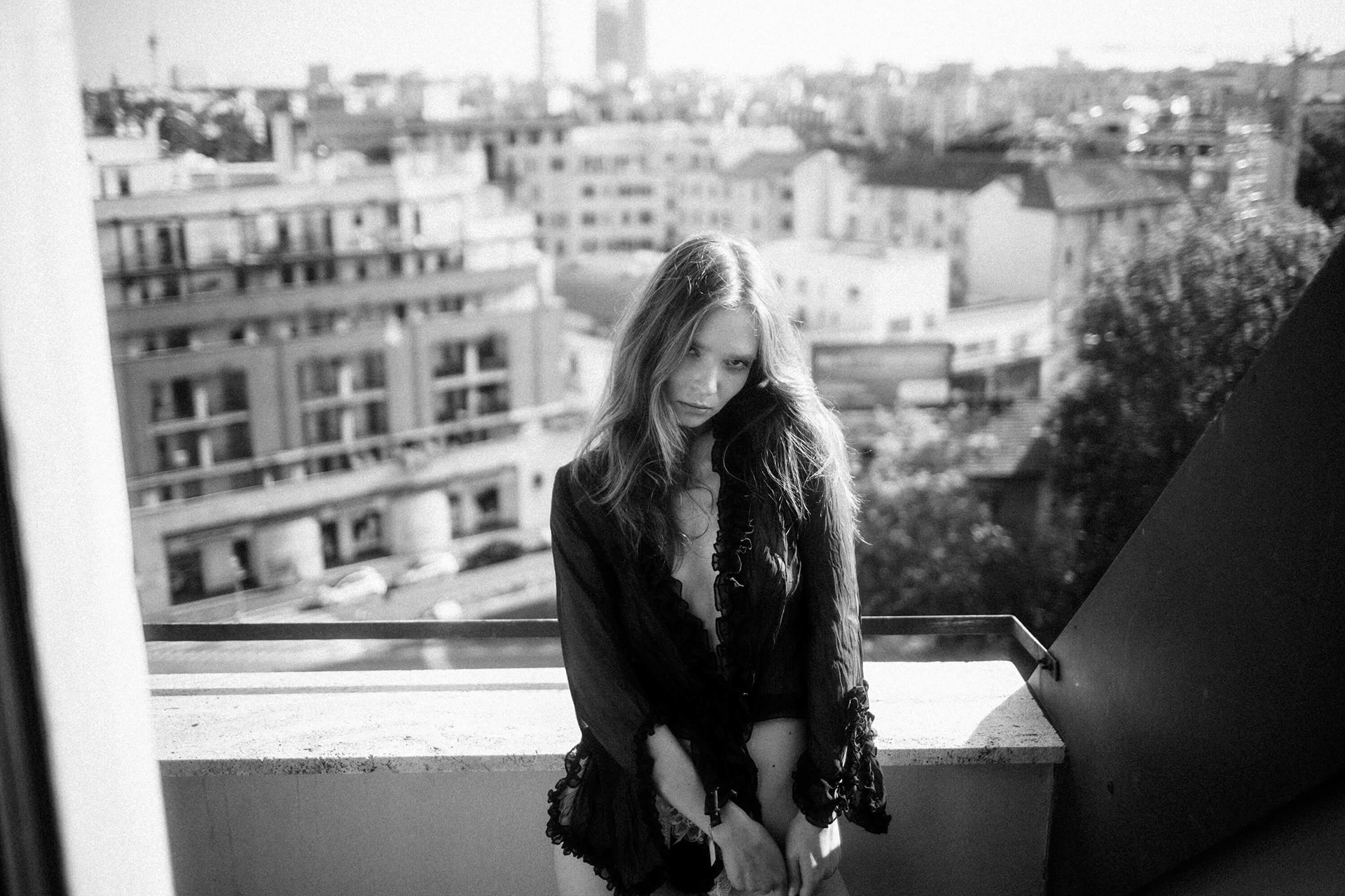 Fine art nude portrait of a woman standing on a balcony, urban landscape softly out of focus in the background