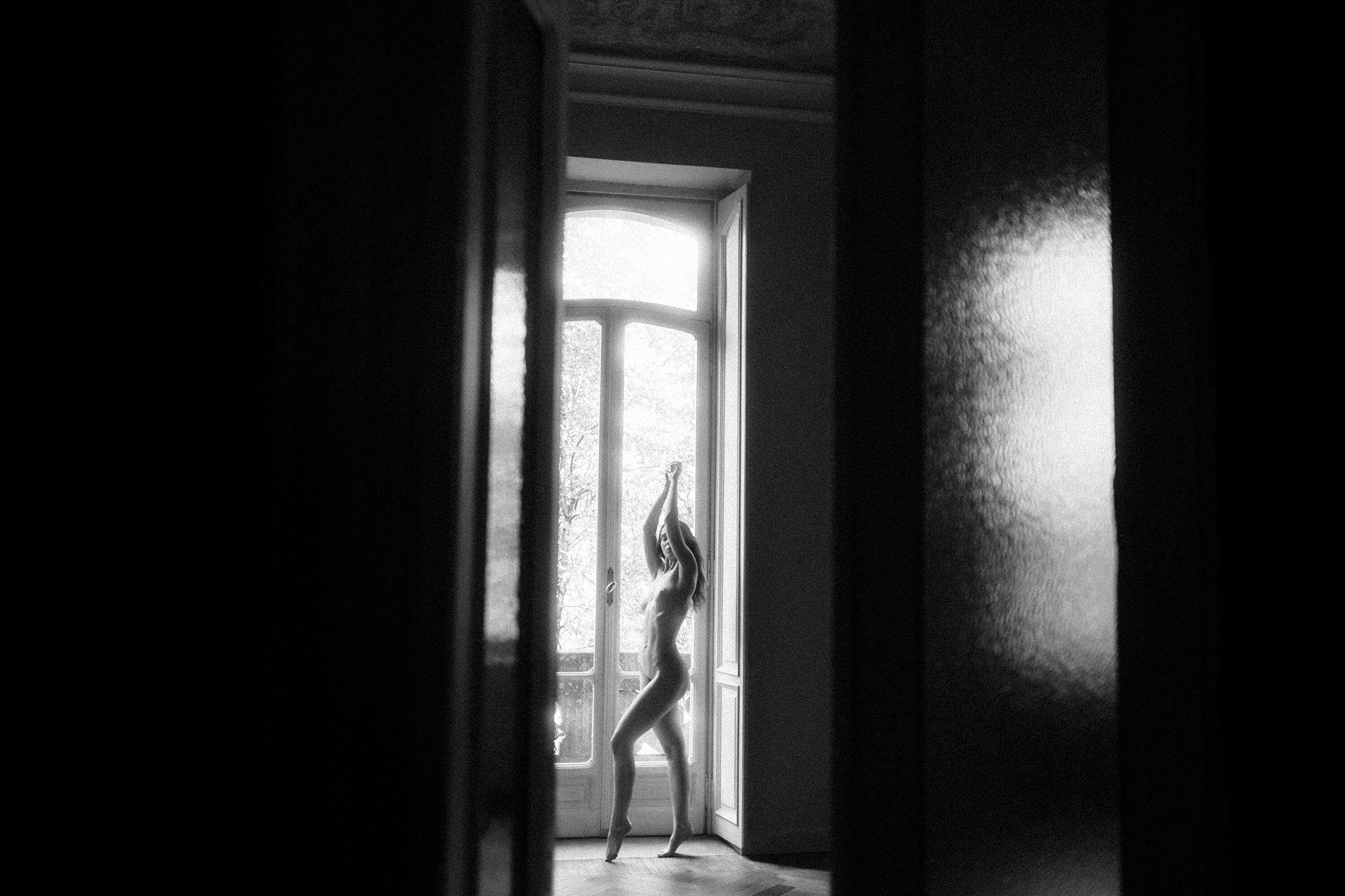 Fine art nude photography of a woman standing with raised arms in a doorway, backlit by natural window light