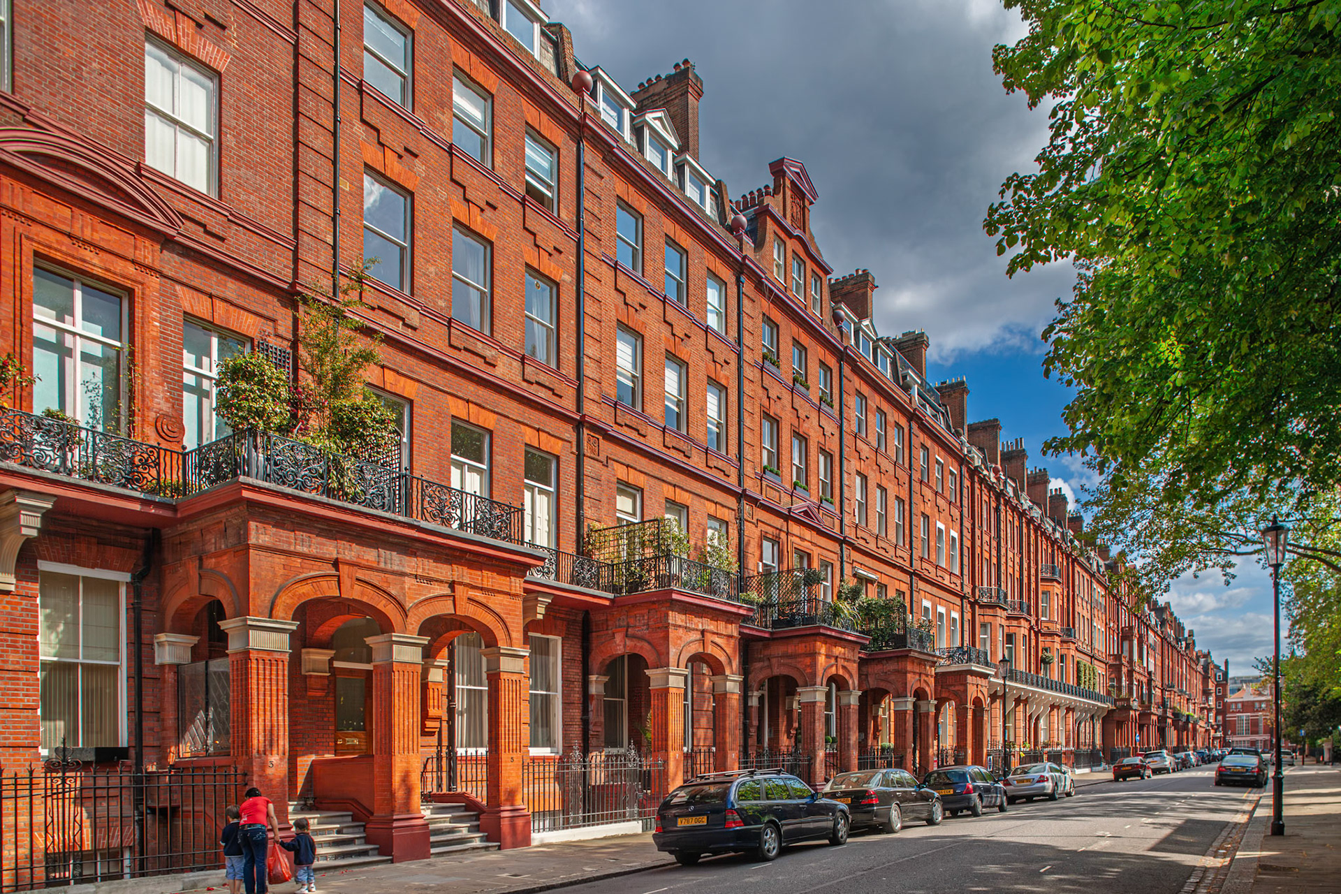Victorian houses in red brick fill the street in Kensington London with colorful details and people walking along the sidewalk on a clear day.