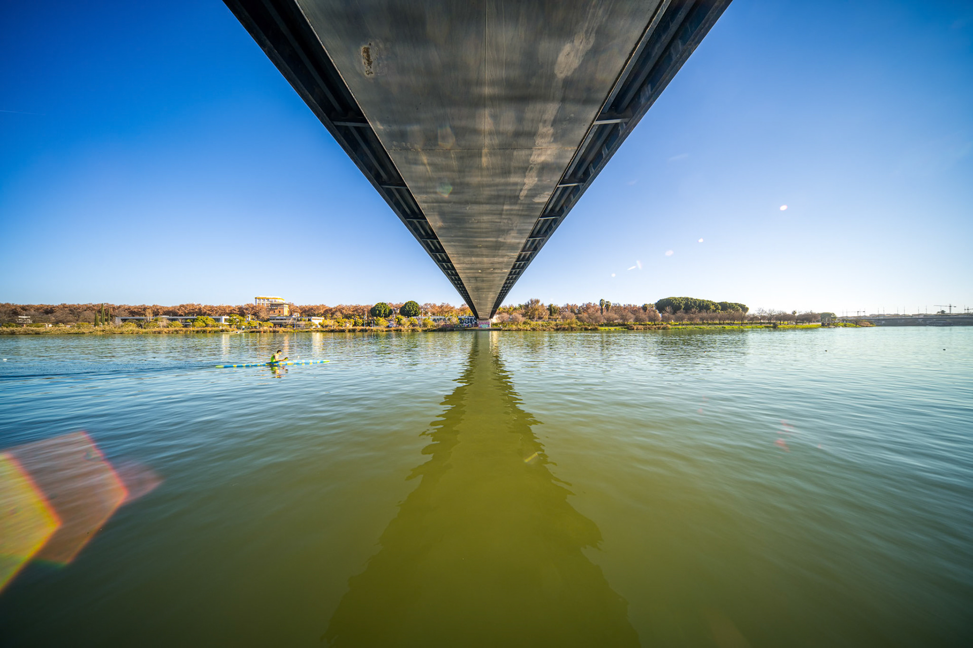 Beneath the Cartuja Footbridge in Seville, steel beams stretch over the Guadalquivir, casting shadows that highlight its elegant design on a clear day.