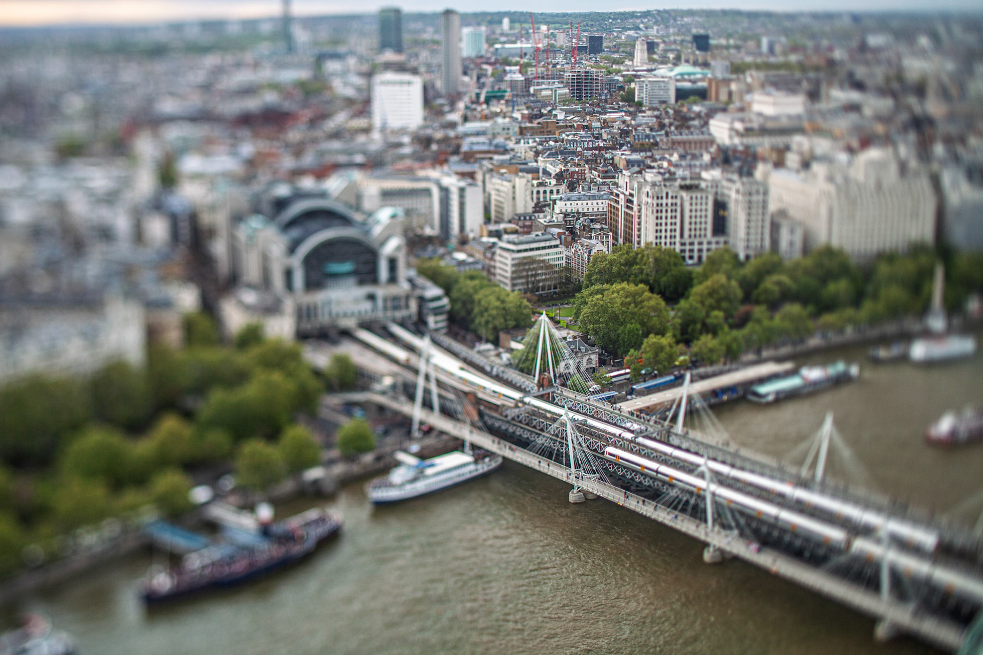 An aerial view shows Hungerford bridge crossing the River Thames near Charing Cross station. The London skyline includes buildings and boats on the water.