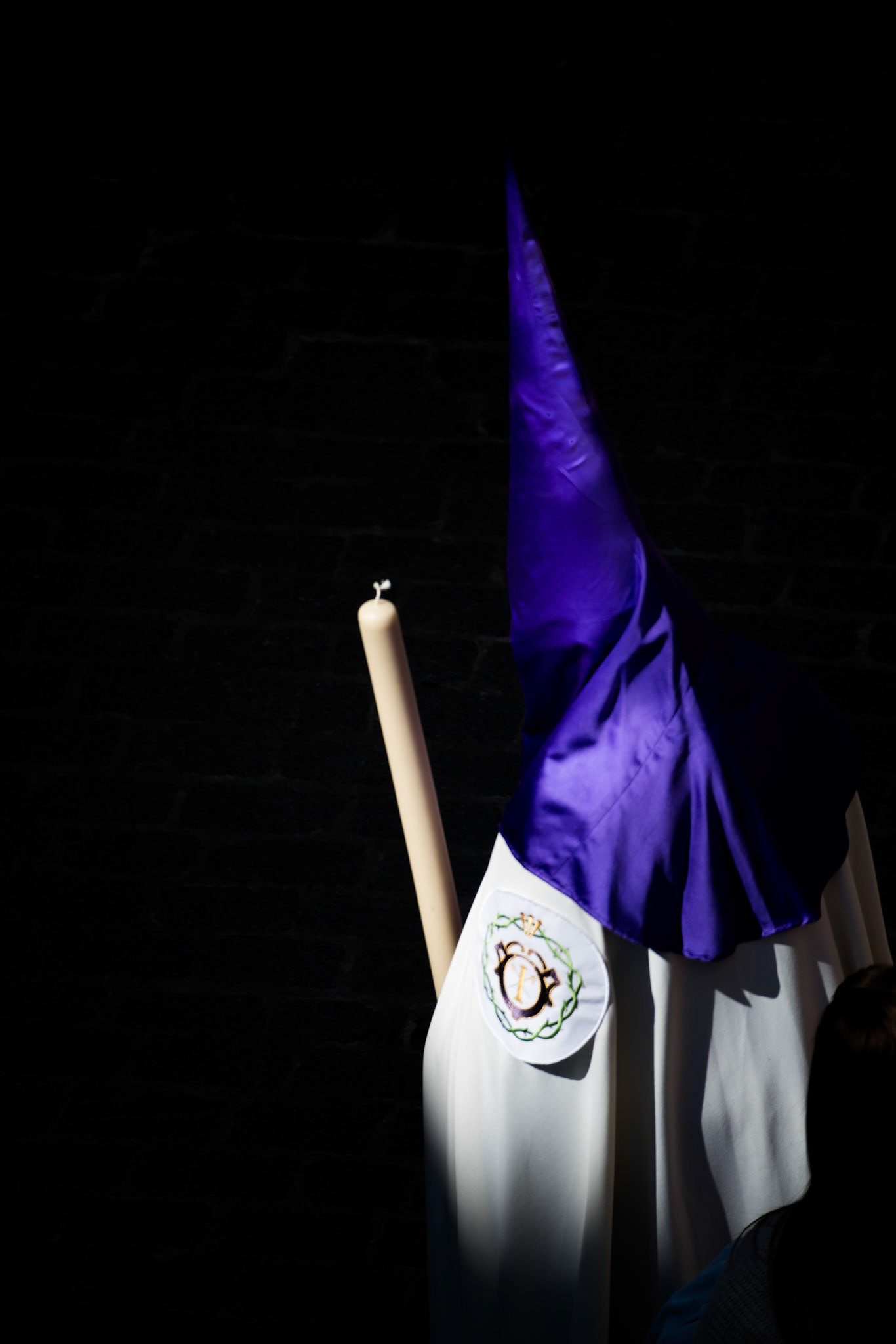 Nazareno stands in bright sunlight during Holy Week in Seville, casting dramatic shadows reminiscent of a tenebrist painting style.