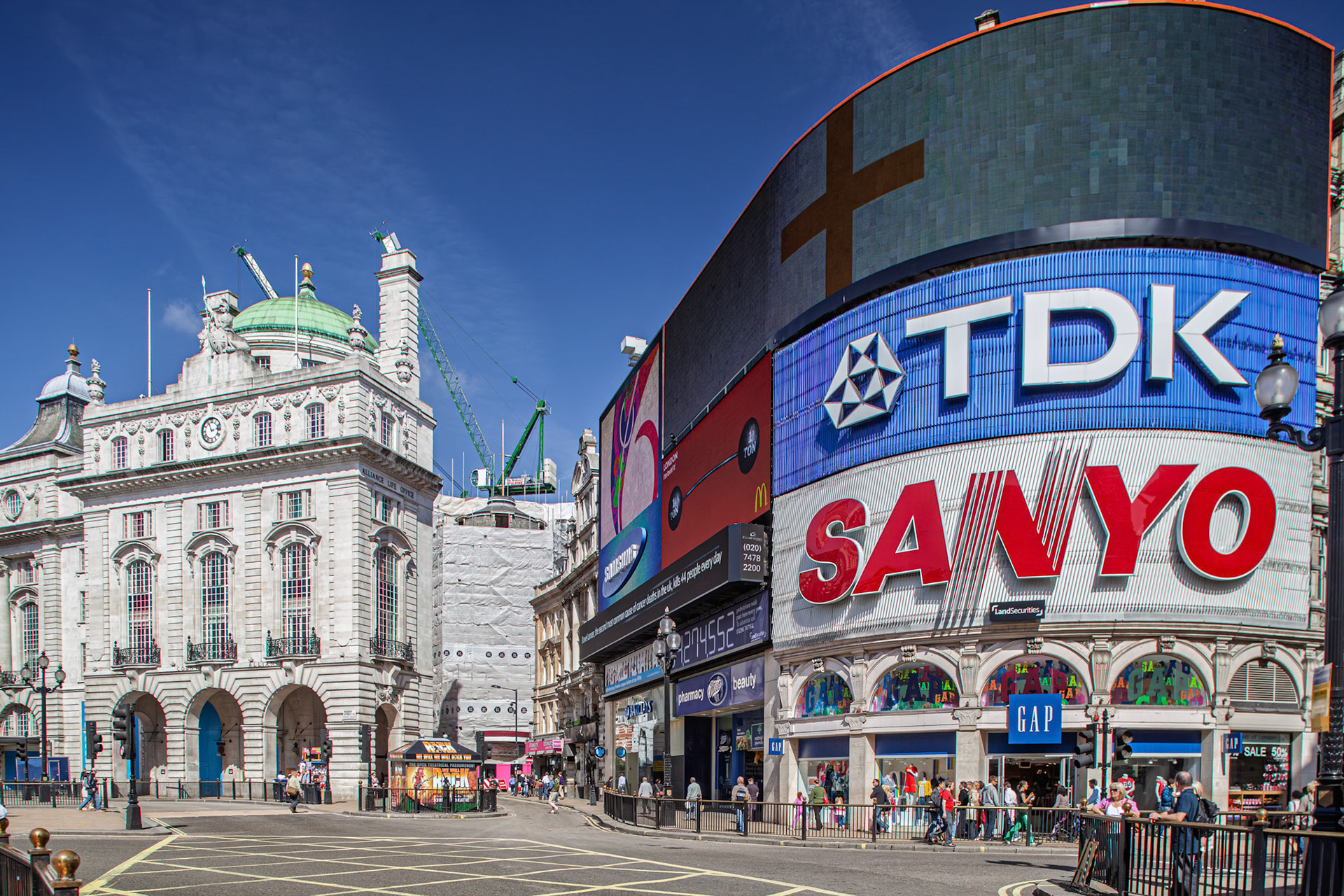People walk through Piccadilly Circus in Westminster London during a clear day. Bright signs and historic buildings surround the area.
