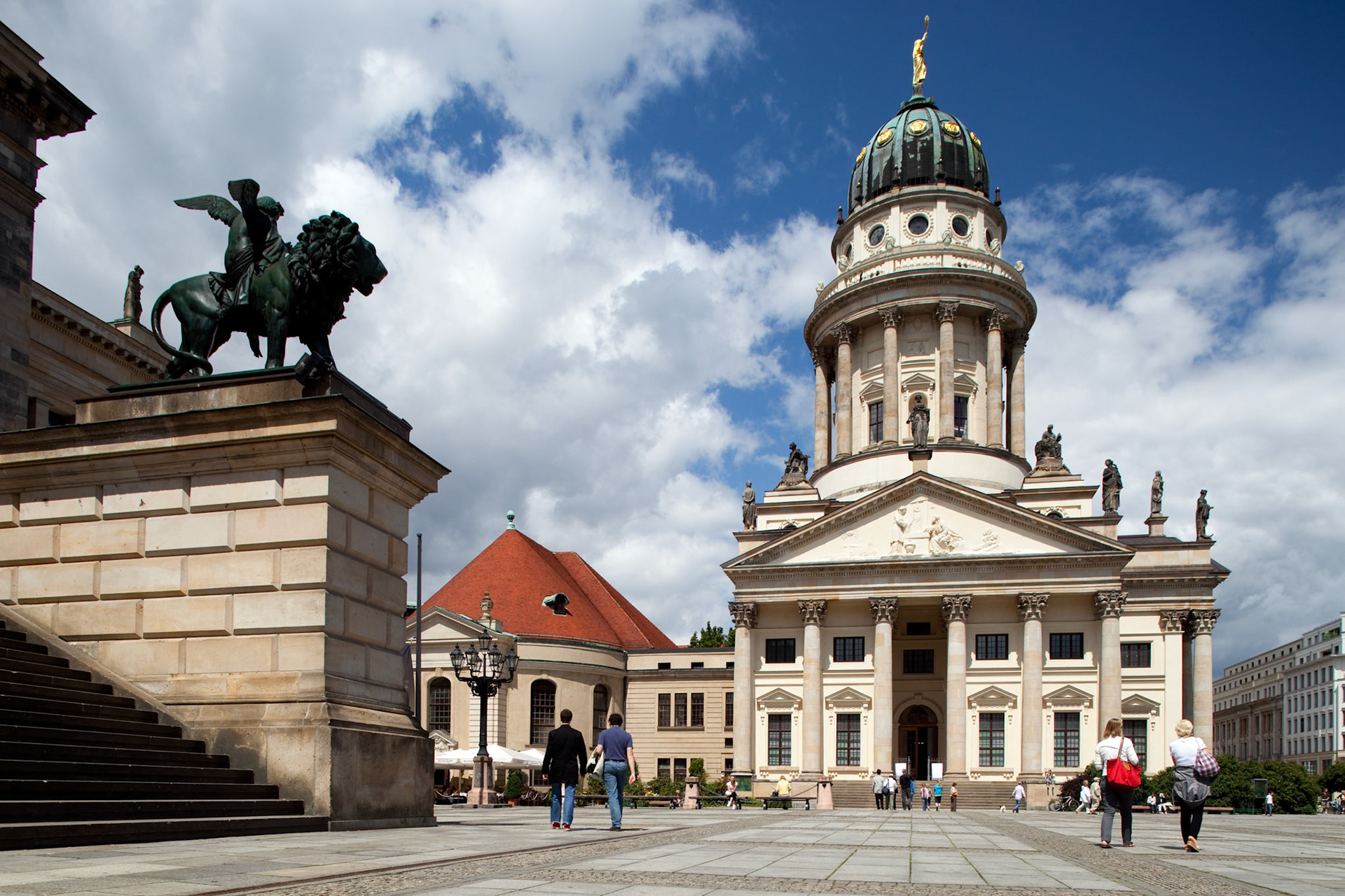People stroll around the stunning Französische Dom at Gendarmenmarkt in Berlin, showcasing its beautiful architecture under a blue sky.