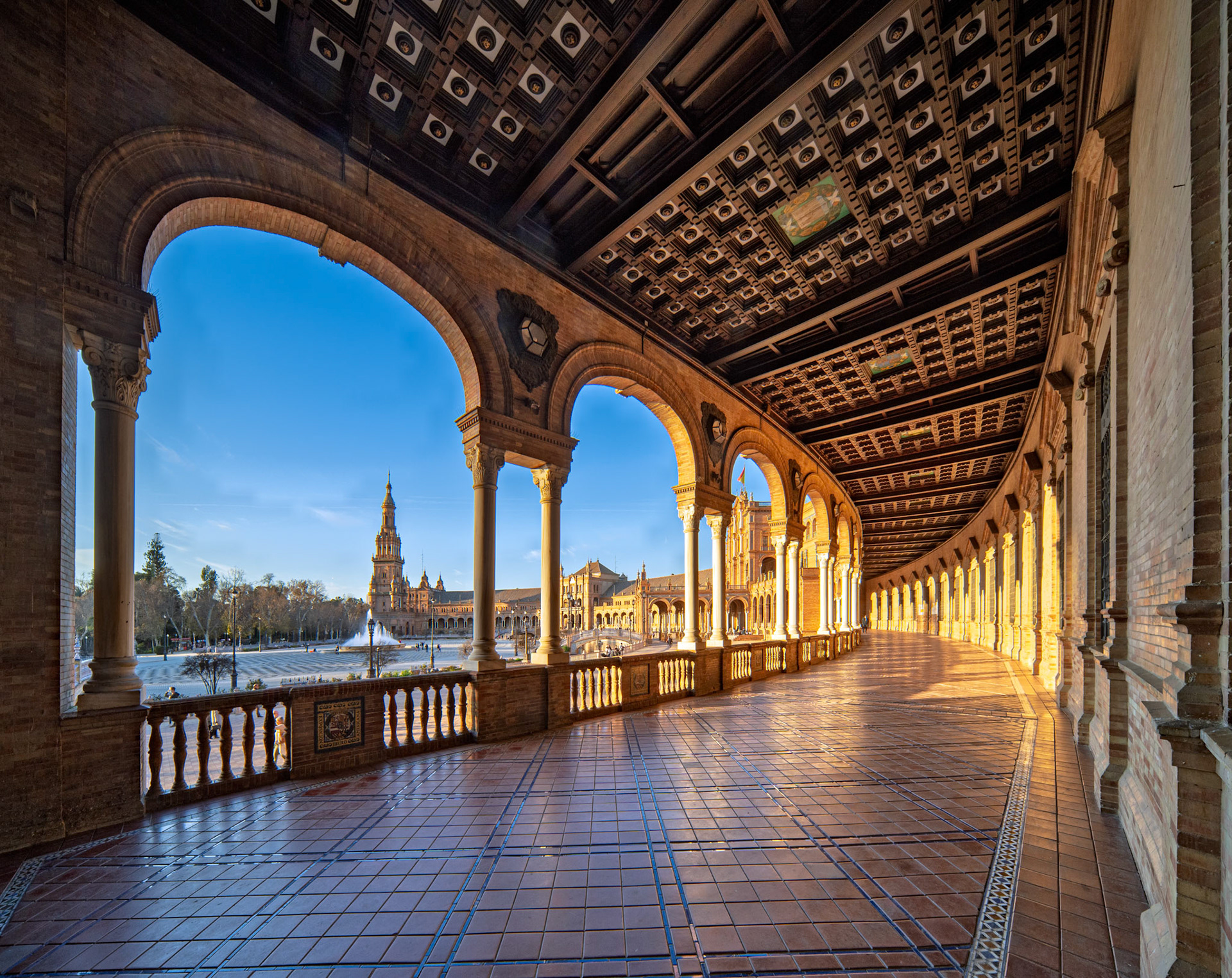 Covered gallery at Plaza de España showcases neomudéjar ceiling with glowing geometric panels and crests, bathed in warm sunlight.