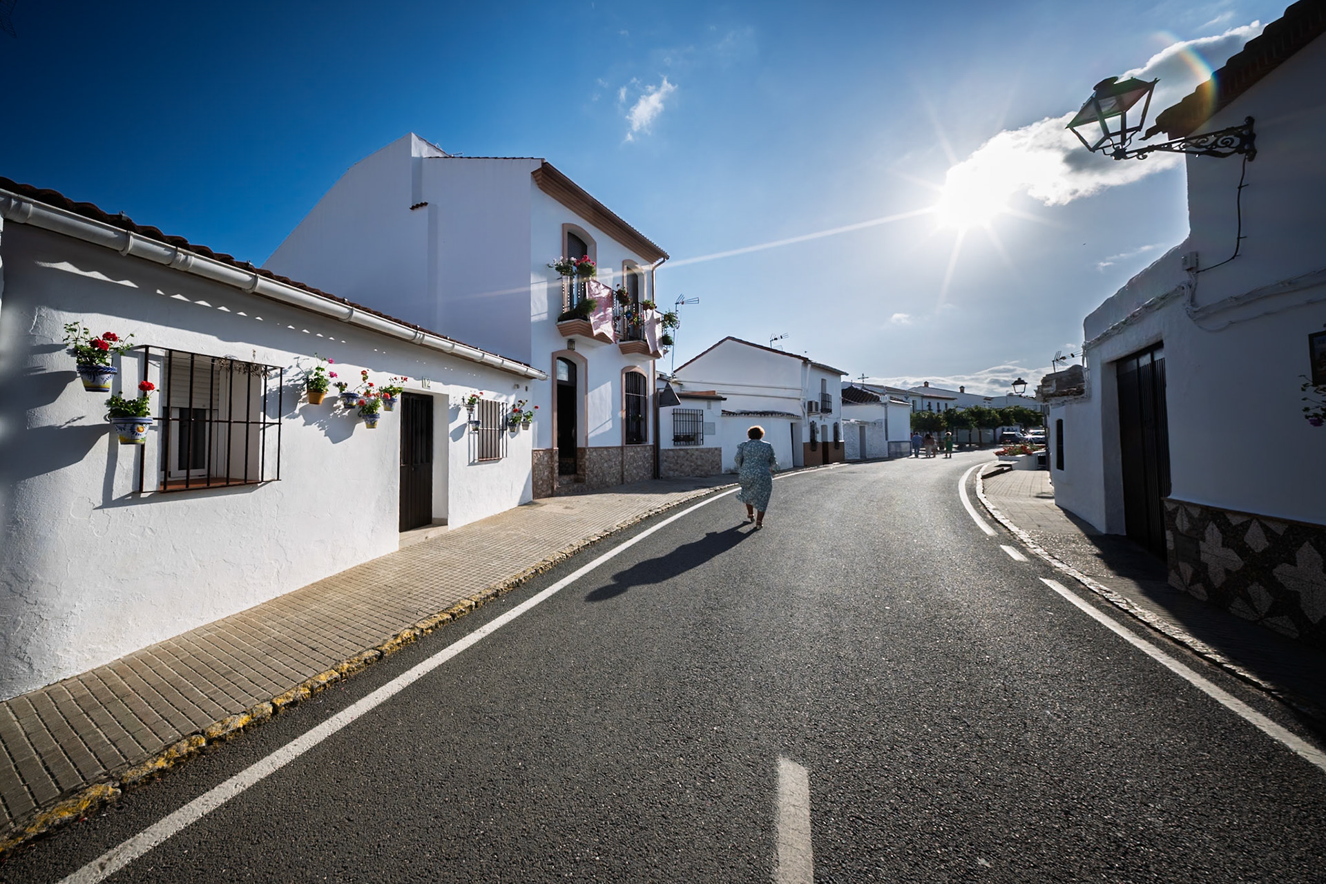 A person walks along a sunlit street in El Madroño, surrounded by whitewashed buildings adorned with flowers during a clear day.