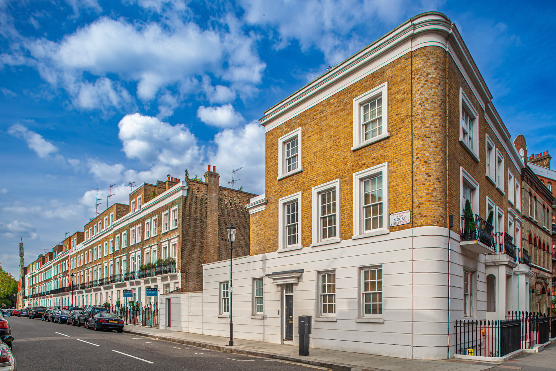 A view of a building on Moore Street in Kensington, London. The sky is blue with clouds, and other buildings are visible along the street.