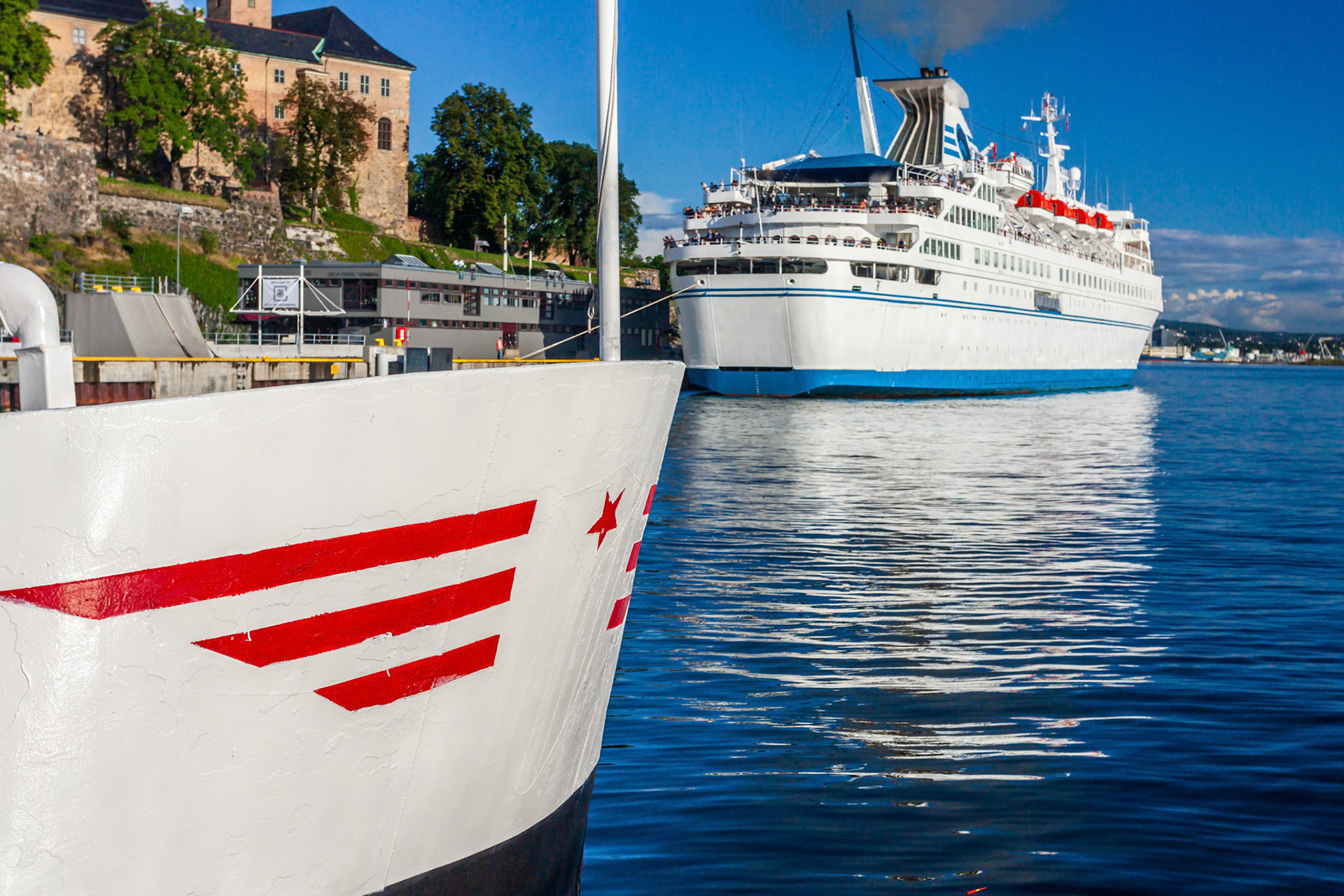 Ships are docked by Akershus fortress in Oslo, Norway, reflecting on the calm waters under a clear blue sky.