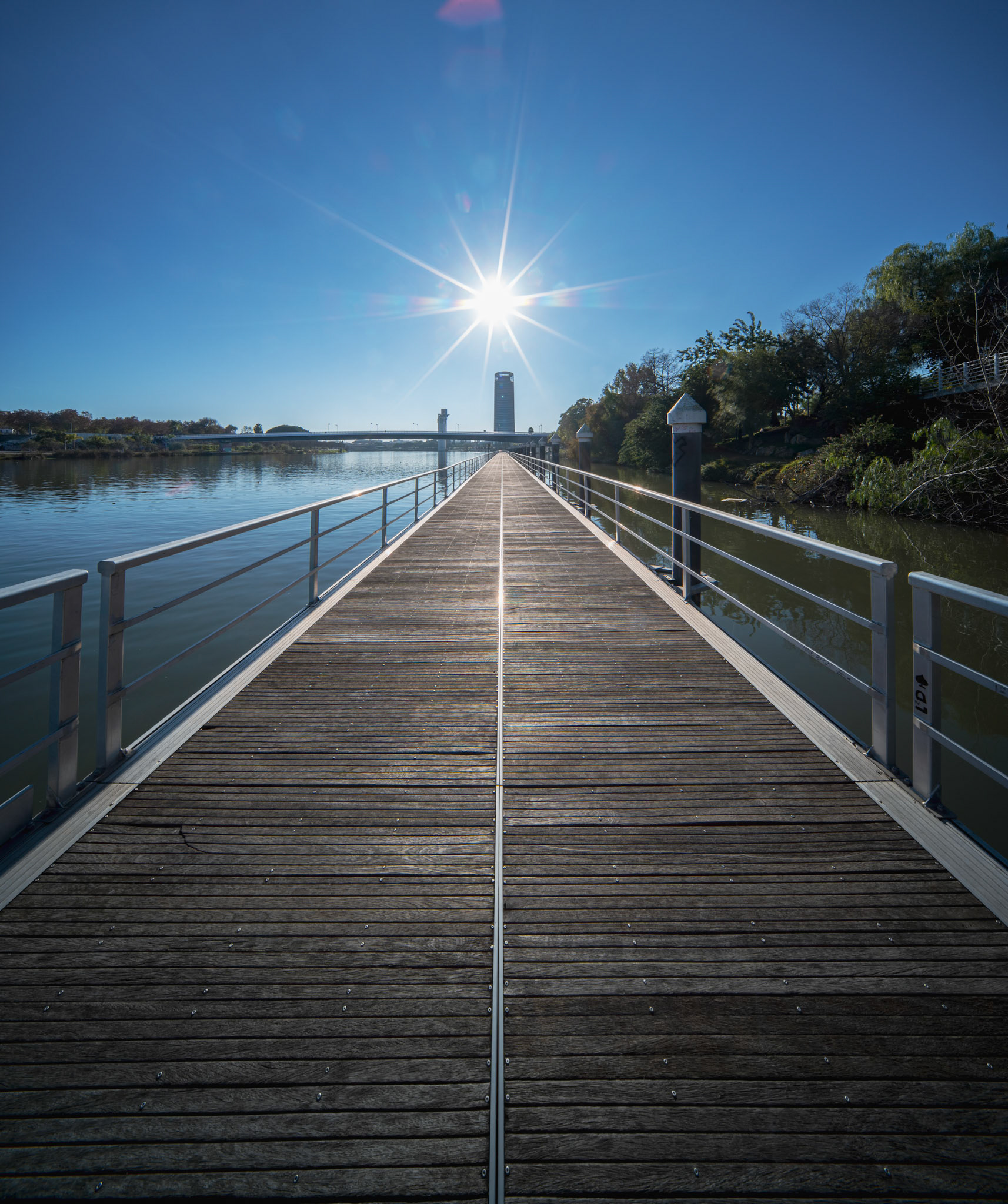 A wooden boardwalk stretches along the Cartuja riverfront in Seville, framed by metal railings and kissed by winter sunlight, revealing serene beauty.