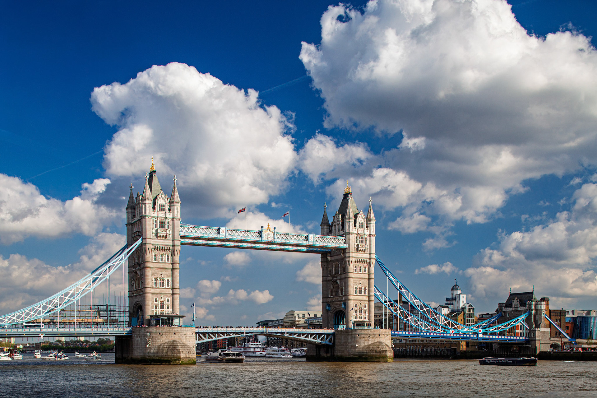 Tower Bridge stands tall over the Thames River as boats pass below on a sunny day in London. Clouds drift in the sky above the historic landmark.
