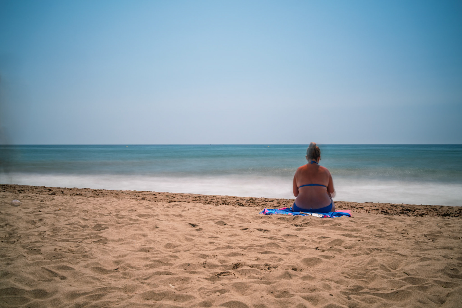 A woman sits peacefully on the sandy beach of Isla Canela, observing the serene sea during a bright daytime long exposure.