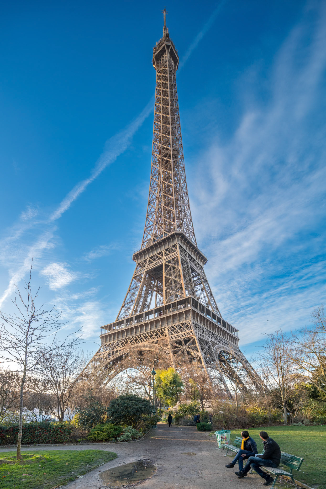 Two men sit on a park bench with the Eiffel Tower looming grandly behind them under an evening sky.