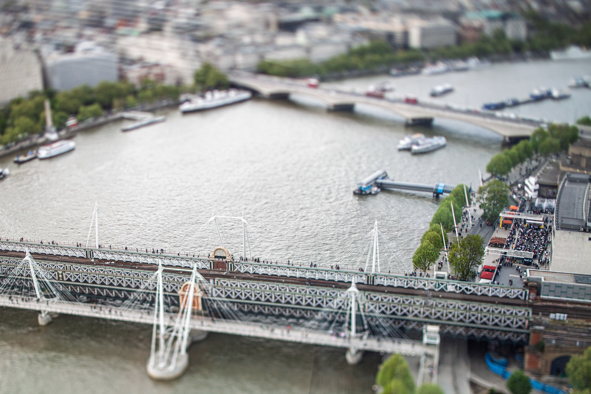 Aerial view shows Hungerford Bridge crossing the Thames with Waterloo Bridge behind. Boats move on the river while people walk on the bridge below.