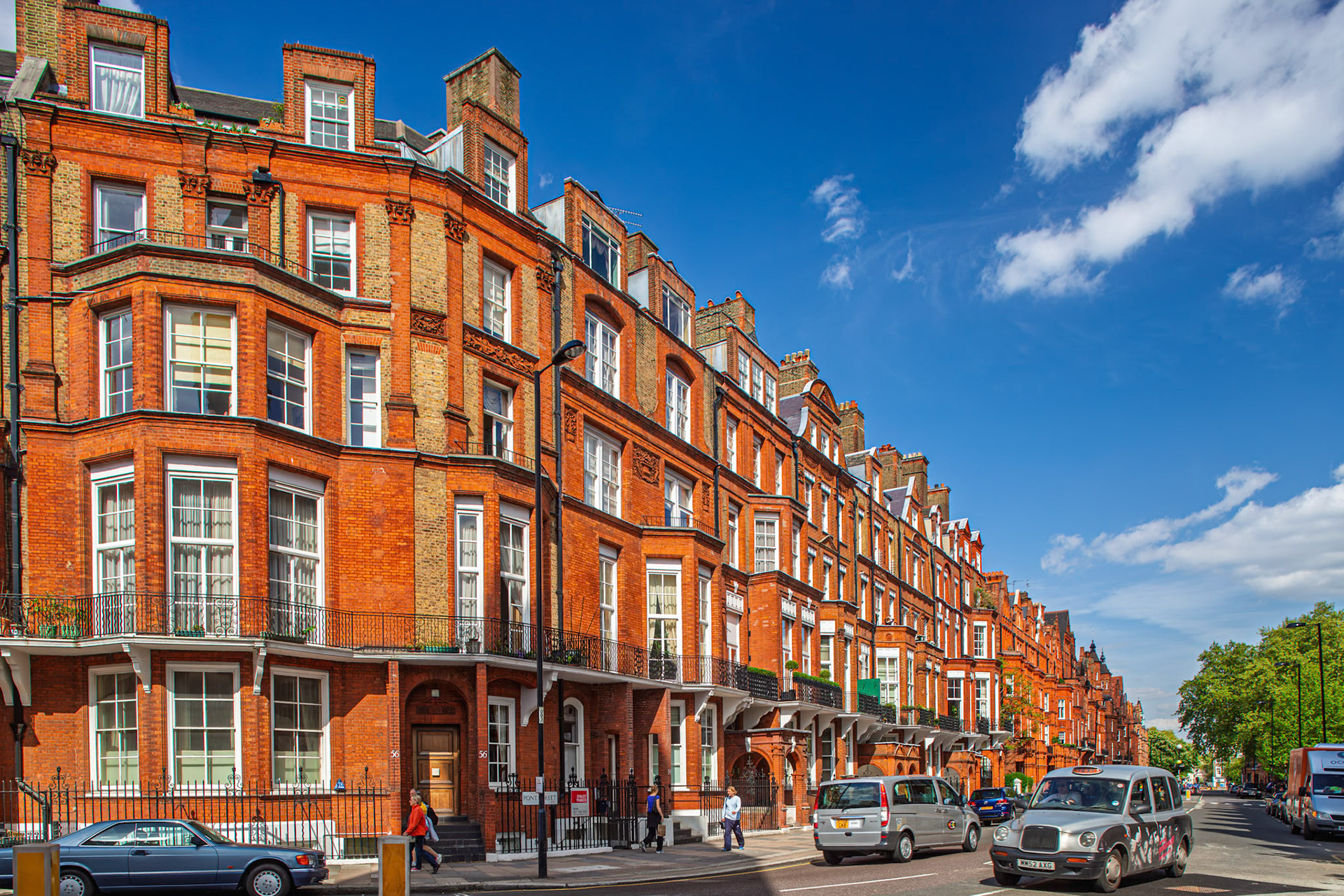 Red brick Victorian houses line Pont Street in Kensington, London. Cars are parked along the street on a sunny day with blue skies.