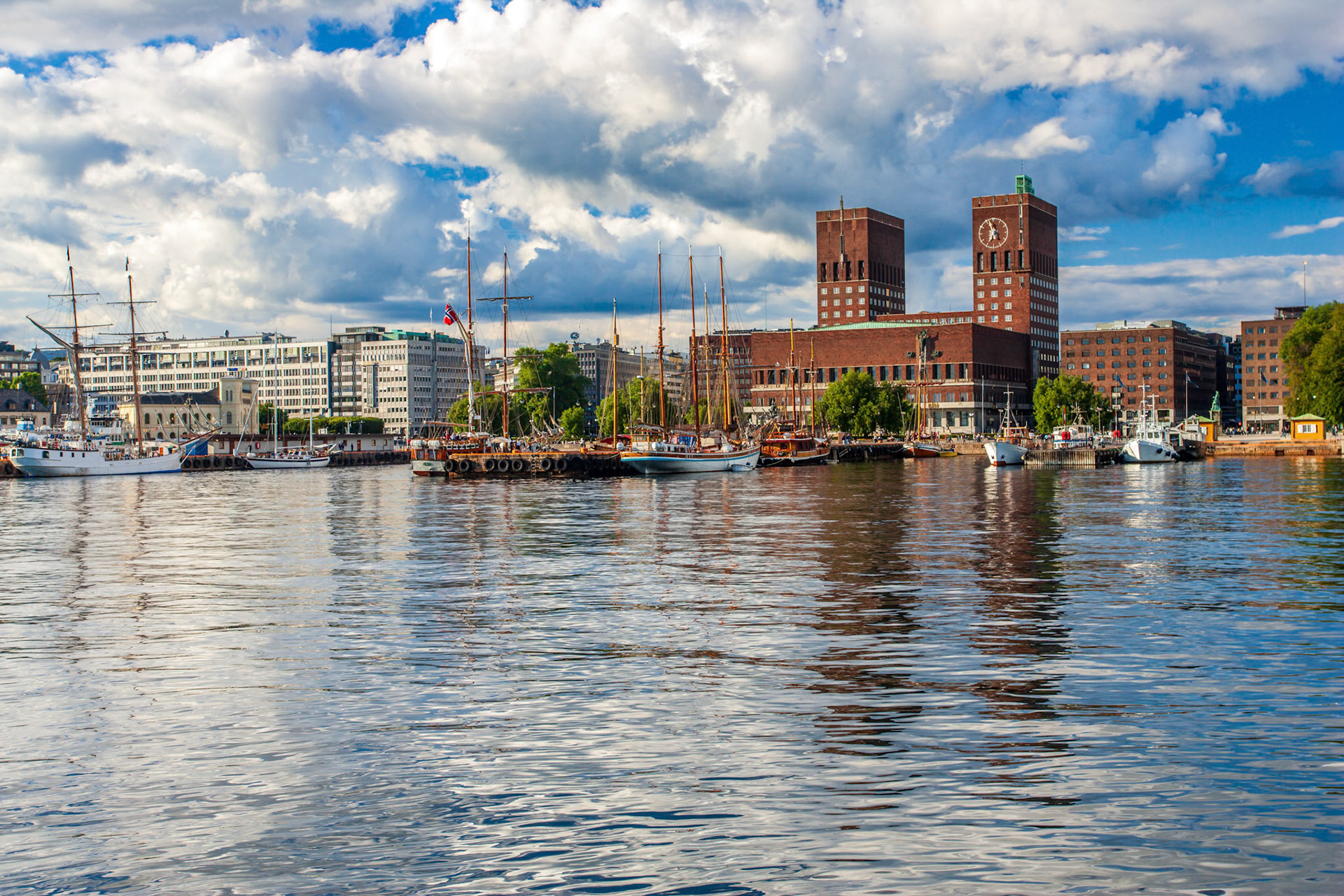 Clear blue skies reflect on the Fjord as Oslo City Hall stands proudly near historical ships and modern buildings.