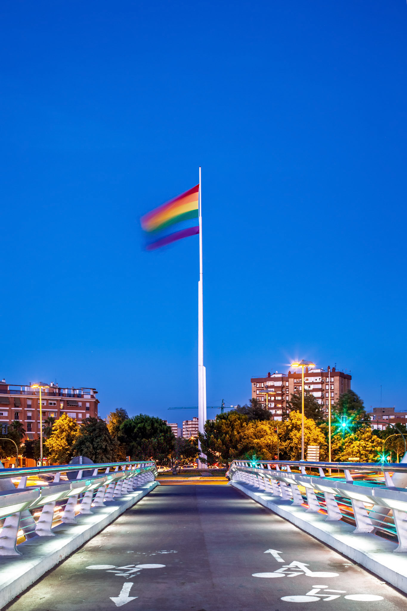 A vibrant rainbow flag stands tall at the Alamillo roundabout in Seville during Pride, reflecting the spirit of diversity as night falls.