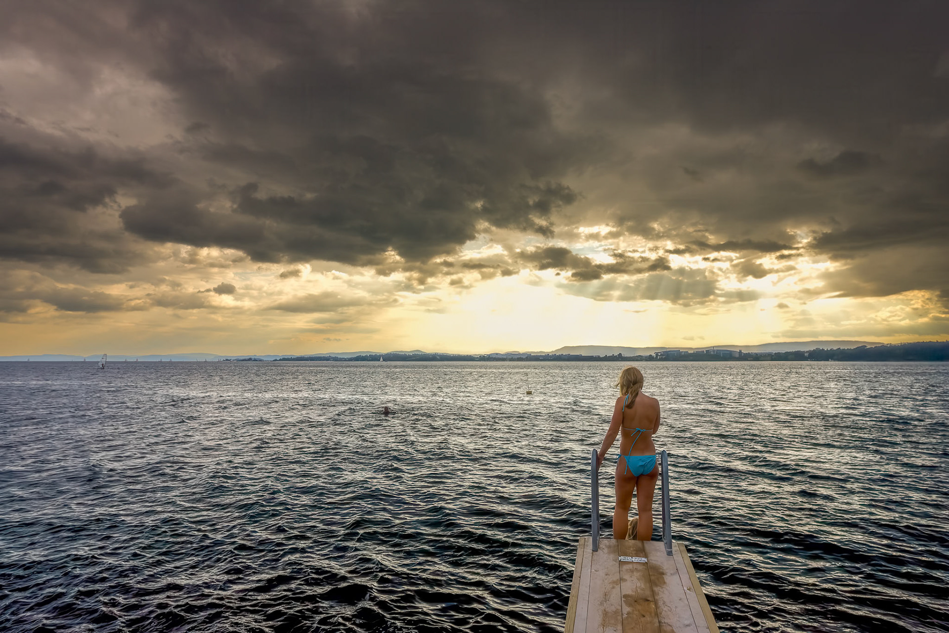 A person stands on a dock, gazing at the calm waters and dramatic clouds during sunset at Paradise Beach in Oslo.